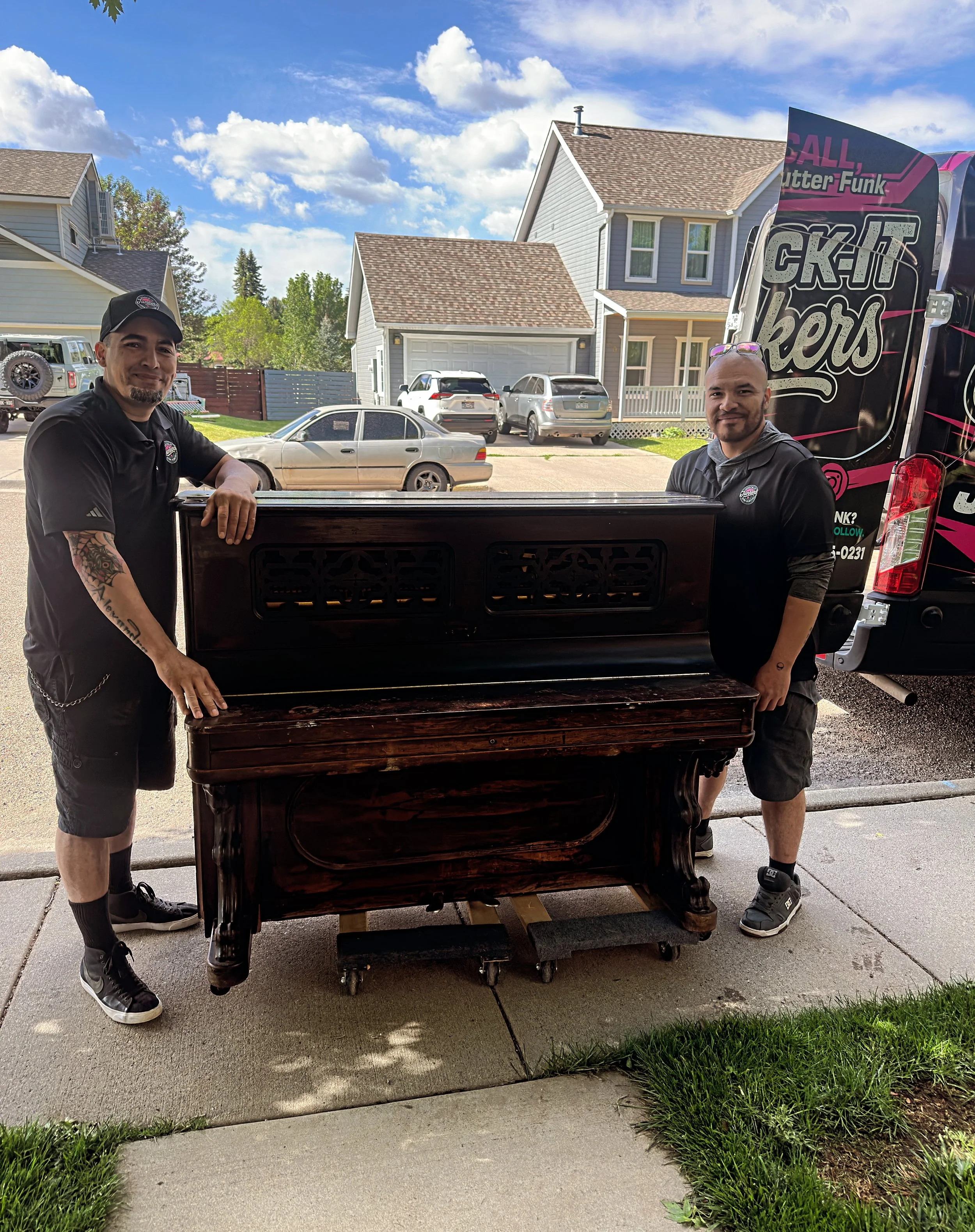 Two men standing outside near a vintage piano and a food truck, with houses and parked cars in the background on a sunny day.