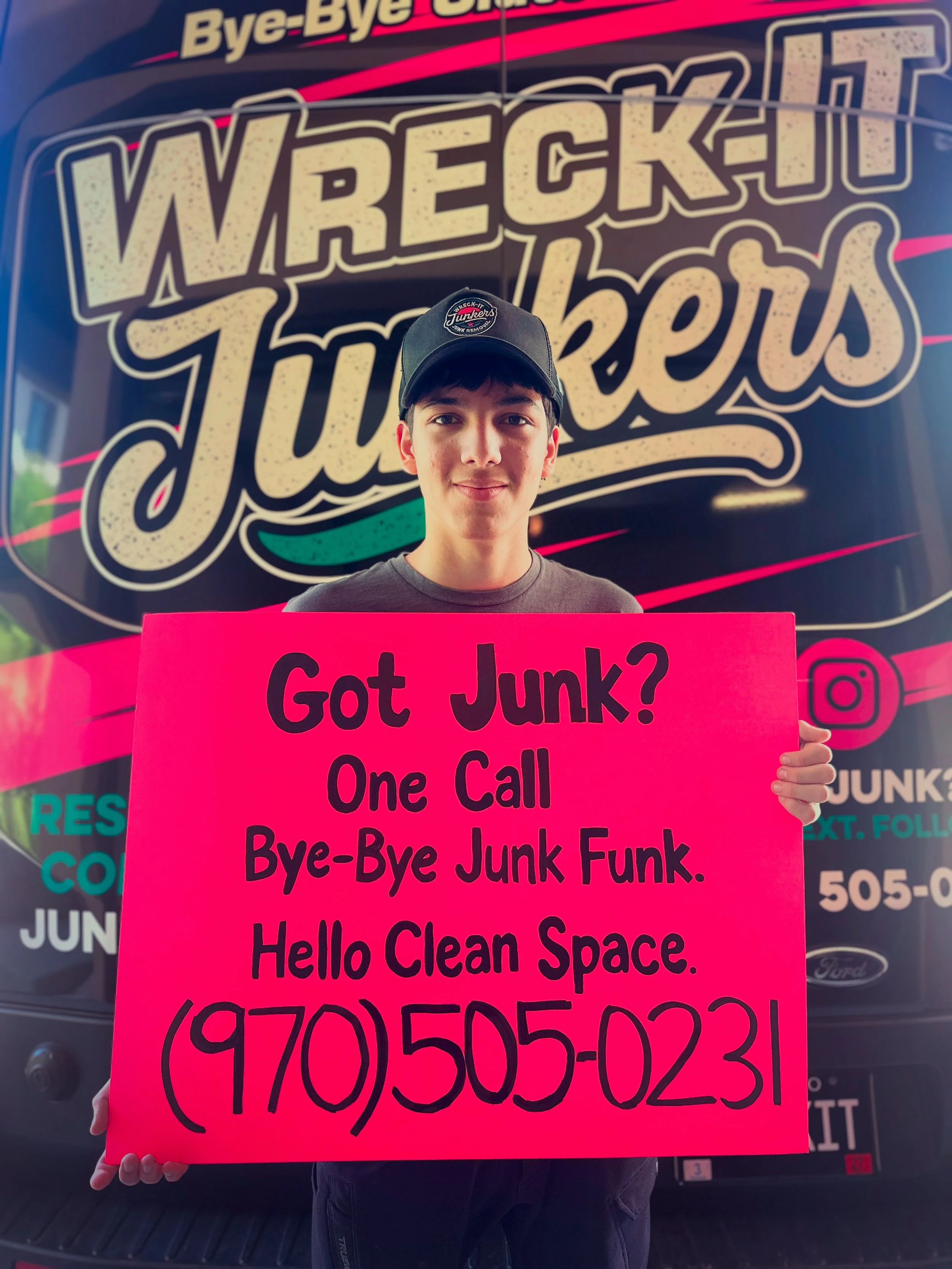 A young person holding a large bright pink sign that reads, 'Got Junk? One Call Bye-Bye Junk Funk. Hello Clean Space,' standing in front of a black truck with colorful branding for a junk removal service.