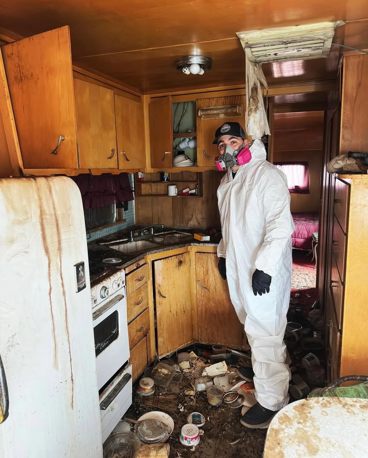 Person wearing protective gear, including a white suit, black gloves, and a respirator mask, standing in a cluttered and damaged kitchen.
