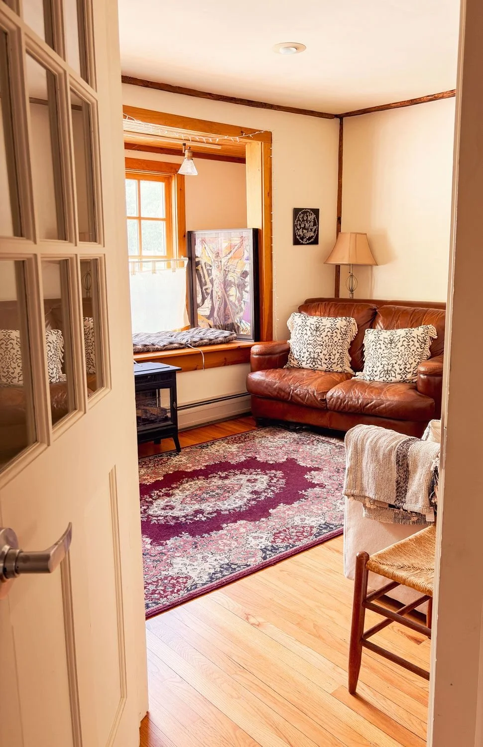 Living room with a brown leather sofa, patterned pillows, a vintage rug, a wooden chair with a striped blanket, a window seat with a cushion, and a window with curtains, decorated in warm tones.