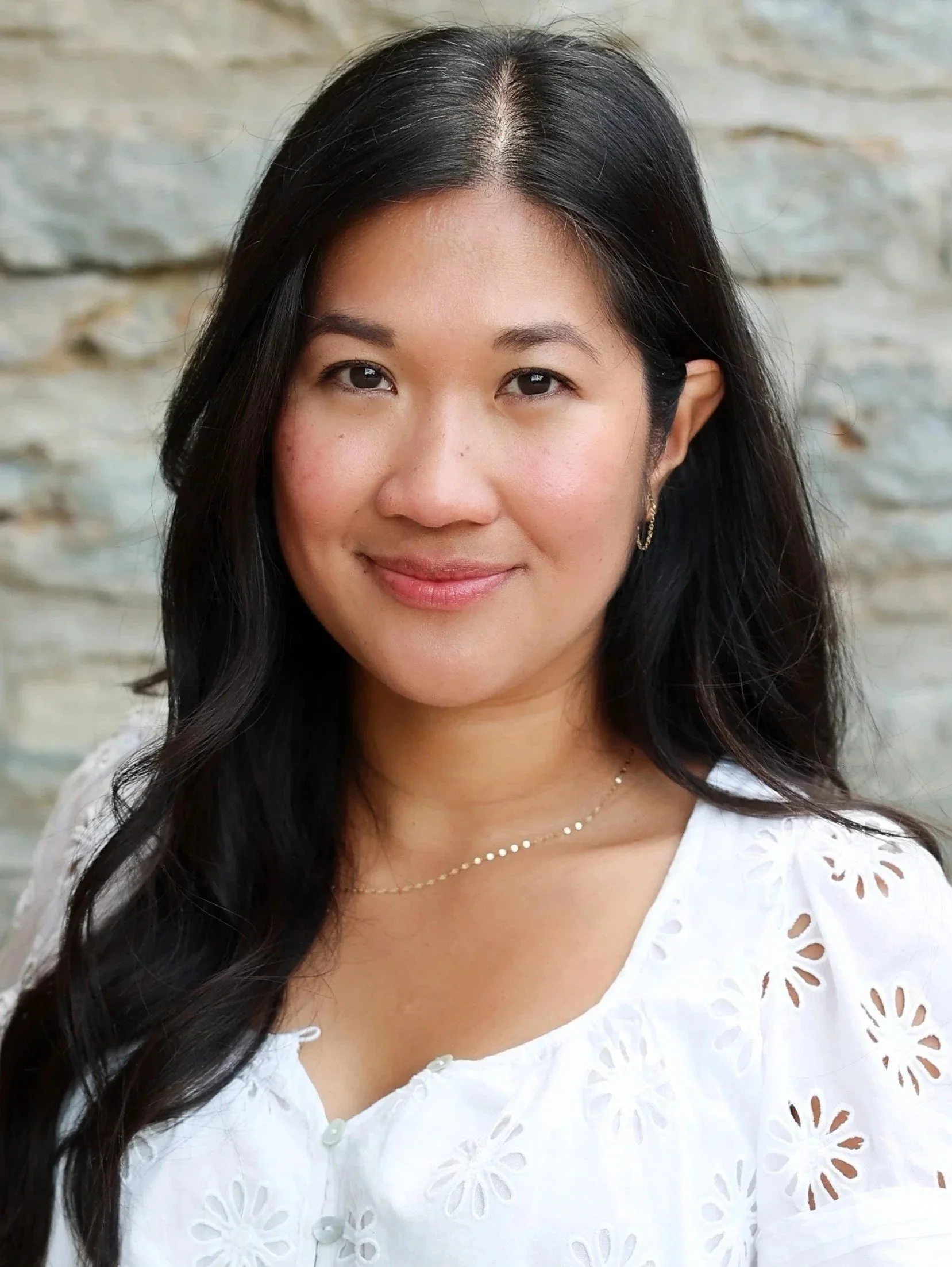A woman with long black hair, wearing a white eyelet embroidered blouse and gold jewelry, smiling against a textured light gray wall background.