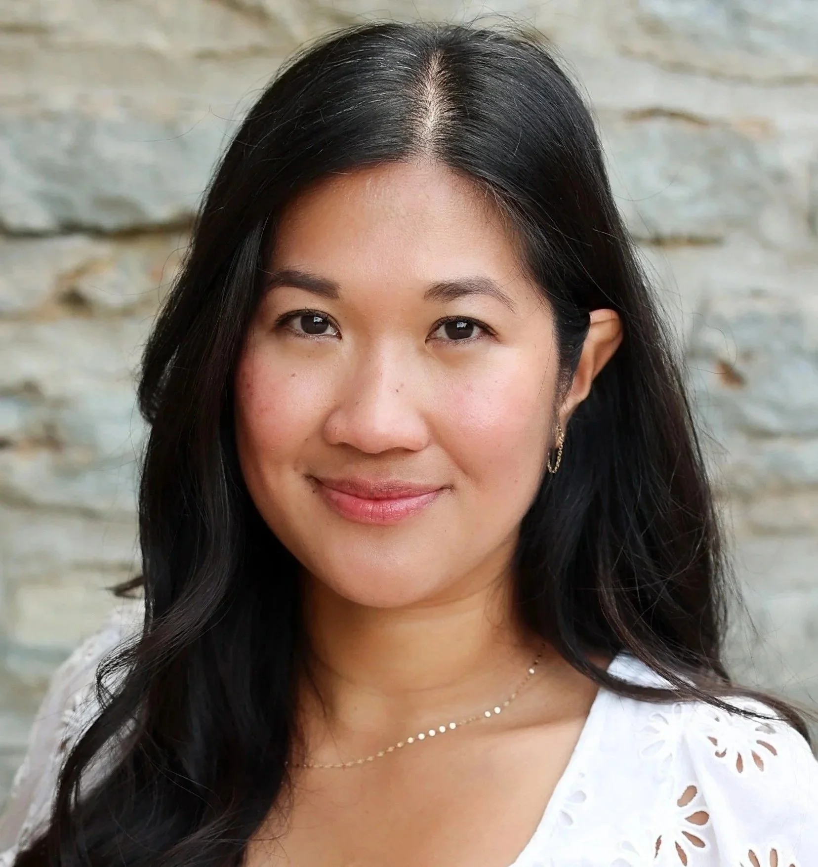 Close-up portrait of a woman with long black hair smiling, wearing a white eyelet blouse and a delicate necklace, with a blurred stone background.