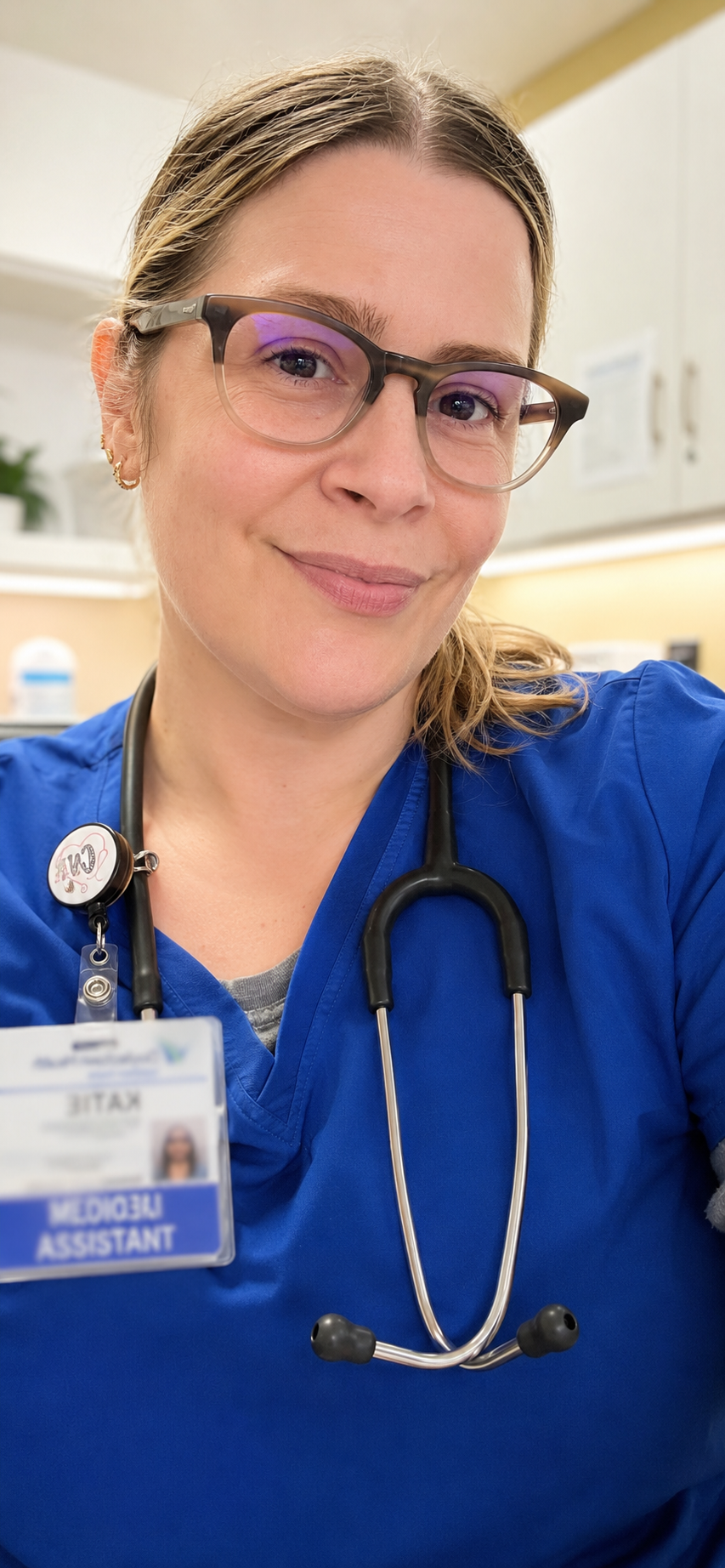 A smiling female healthcare professional wearing glasses and blue scrubs, with a stethoscope around her neck, in a medical setting.
