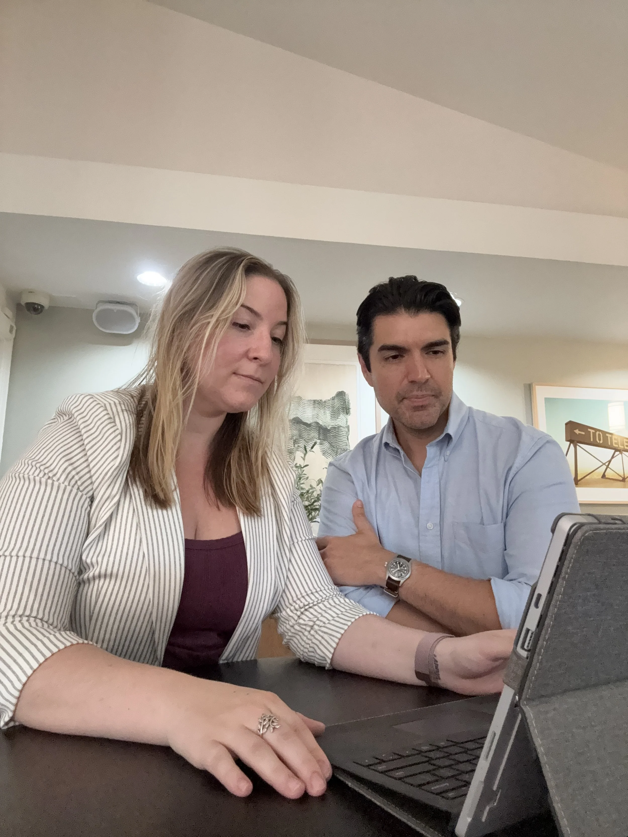 A woman with long blonde hair and a man with dark hair sit at a table looking at a laptop screen. The woman wears a striped blazer and a maroon top, and the man wears a light blue shirt and a watch. They appear to be focused on the laptop, which they are using together in a room with framed artwork on the wall and a sign that reads 'to TELL' in the background.