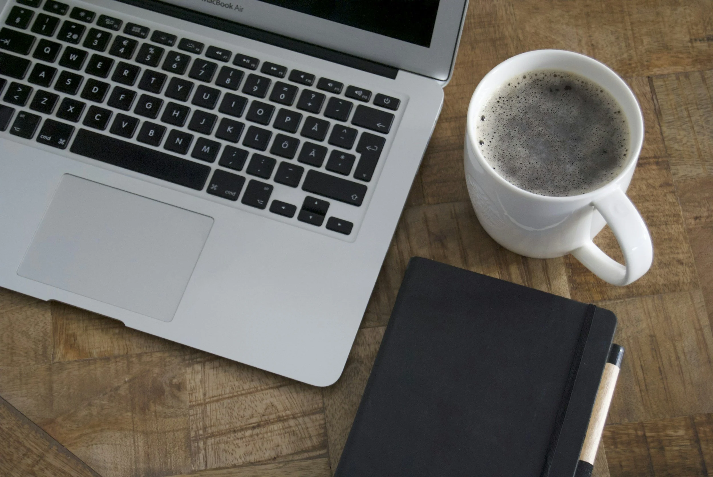 Top view of a wooden table with a silver laptop, a white coffee mug filled with dark coffee, a closed black notebook, and a black pen.