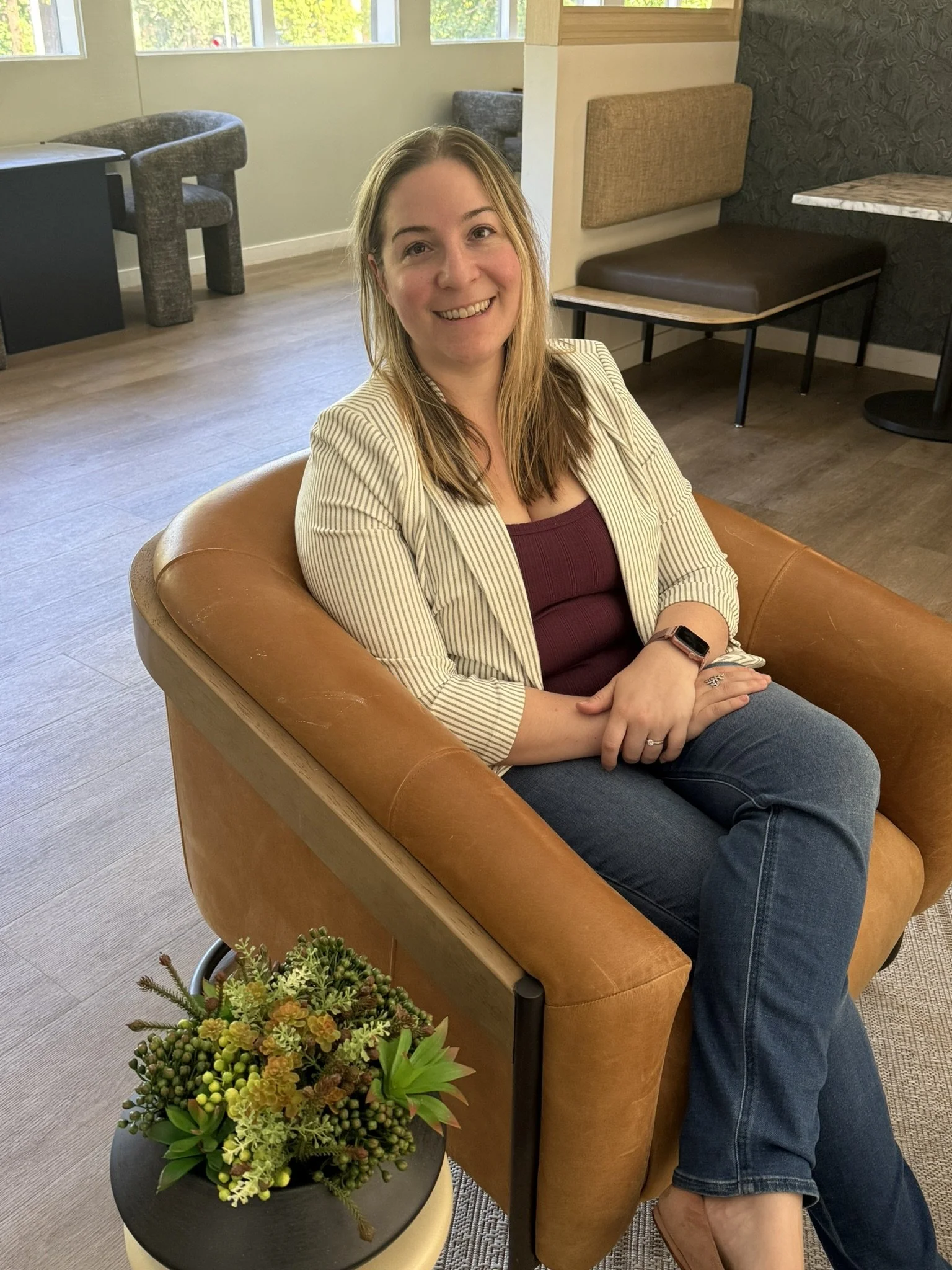 A woman sitting on a brown leather armchair in a cozy, modern room with large windows, a side table with a potted plant, and a patterned accent wall.