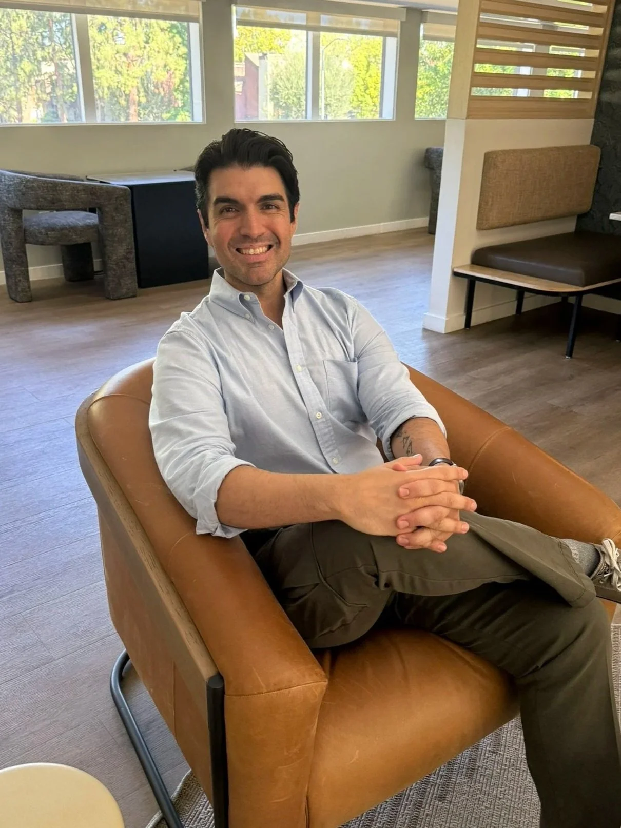A man with dark hair and a light blue button-up shirt sitting on a tan leather armchair, smiling at the camera, in a bright room with large windows and wooden flooring.