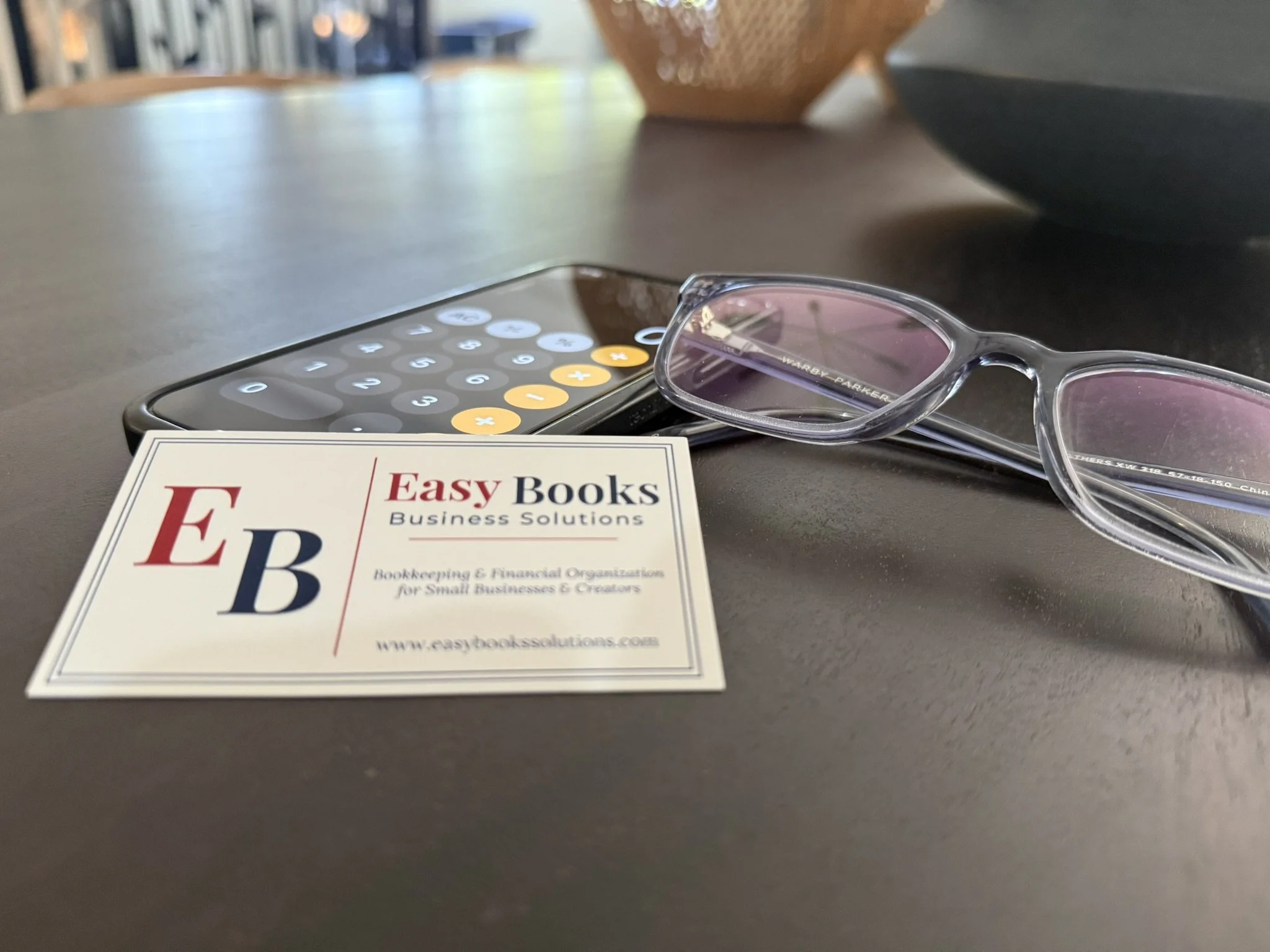 Close-up of eyeglasses, a smartphone with a calculator app open, an 'Easy Books' business solutions pamphlet, and a small beige basket on a table surface.