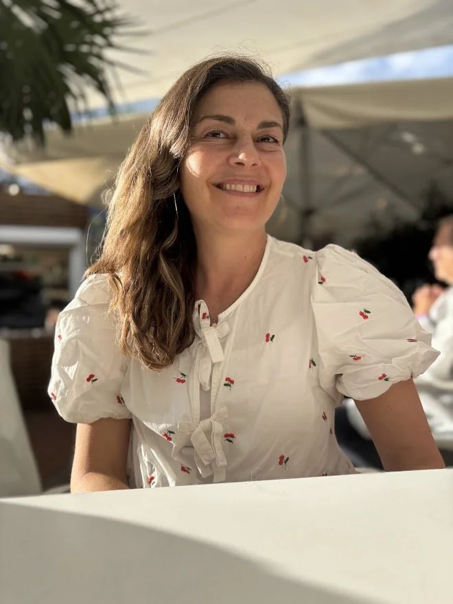 A woman with long wavy brown hair smiling at a camera, wearing a white blouse with small cherry patterns and puffy short sleeves, sitting outdoors under a canopy with sunlight.