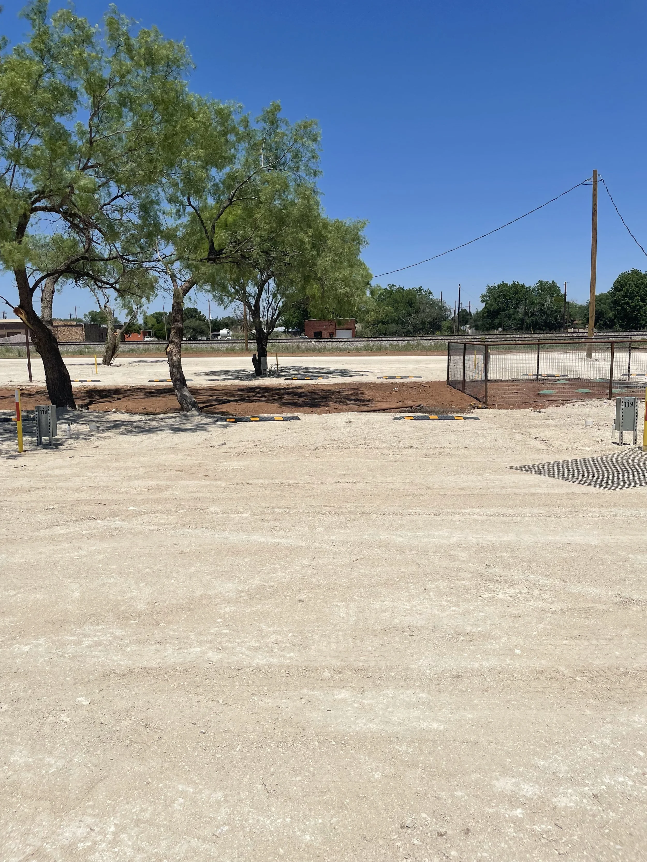 Empty parking lot with trees, a chain-link fence, and utility poles under a clear blue sky.