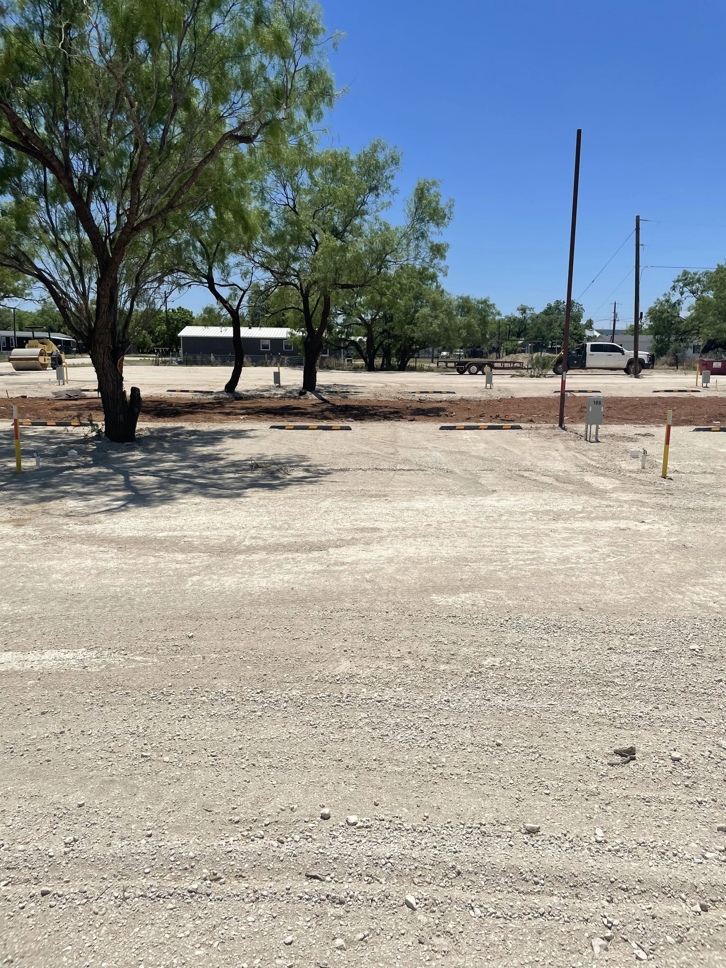 A mostly empty parking lot with trees, utility poles, a small building, and an orange-colored construction roller in the background under a clear blue sky.