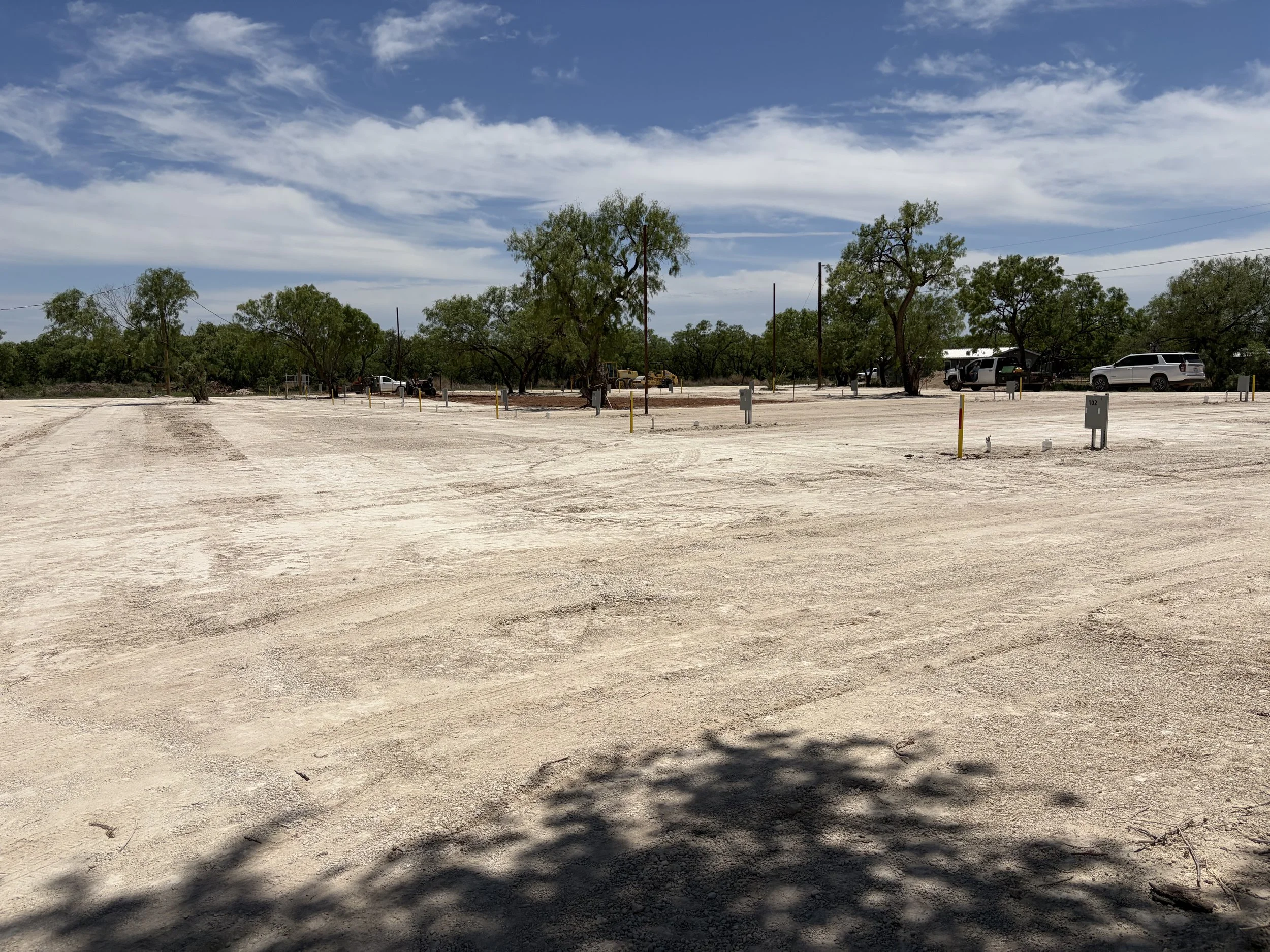 Empty parking lot with trees, utility poles, and a few vehicles under a partly cloudy sky.