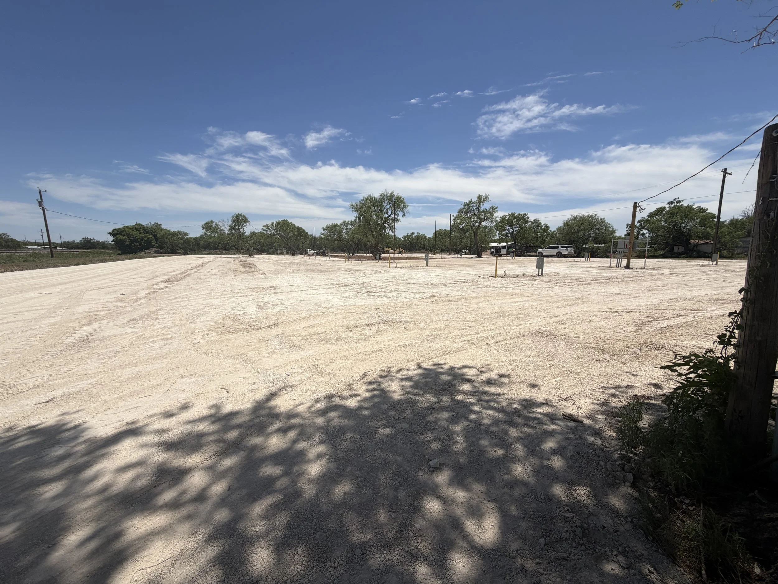 A wide, open dirt parking lot under a blue sky with wispy clouds. There are trees and some vehicles in the distance, with utility poles along the perimeter.
