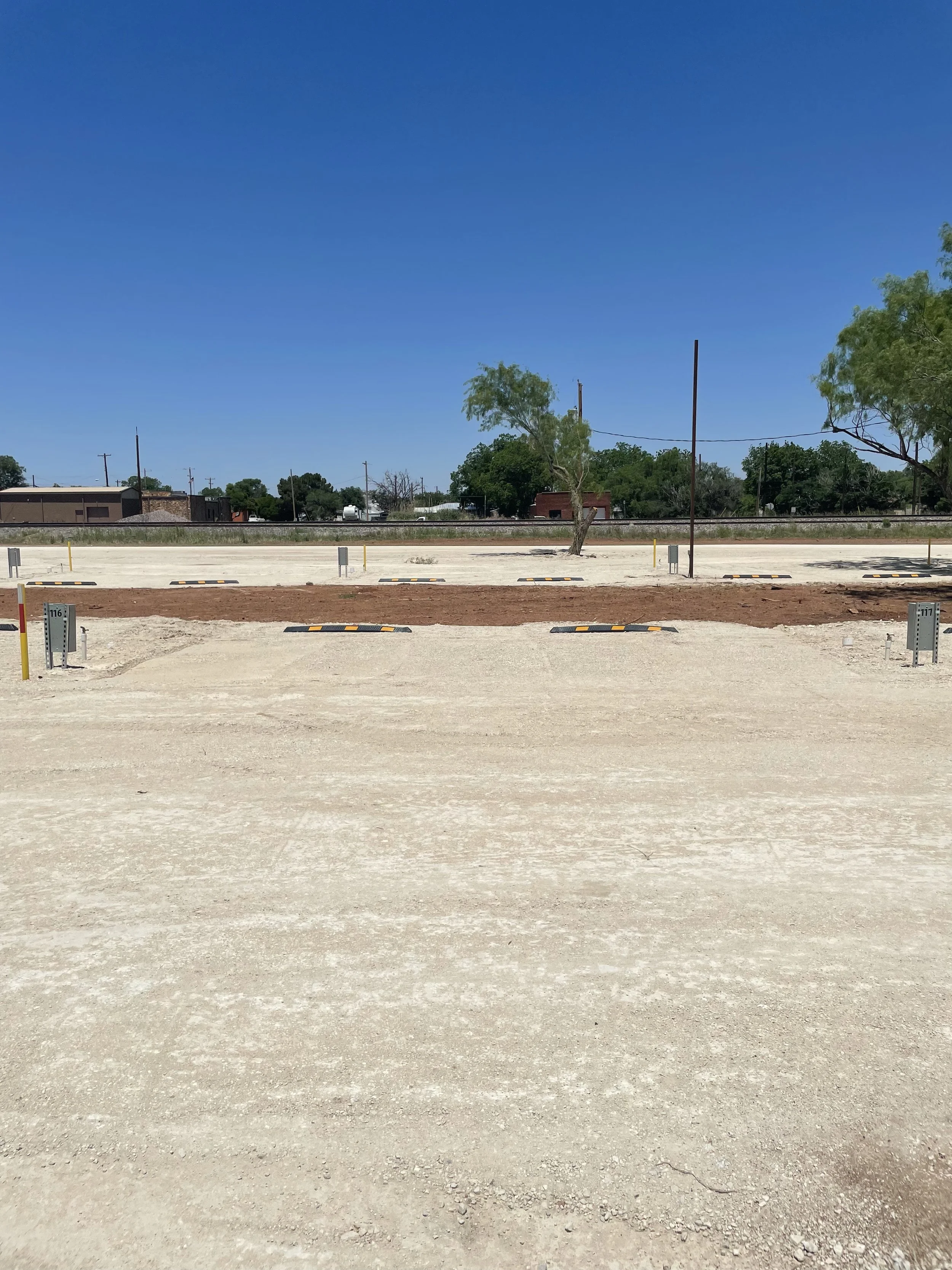 A parking lot with marked parking spaces, utility boxes, trees, and a railway track in the background under a clear blue sky.