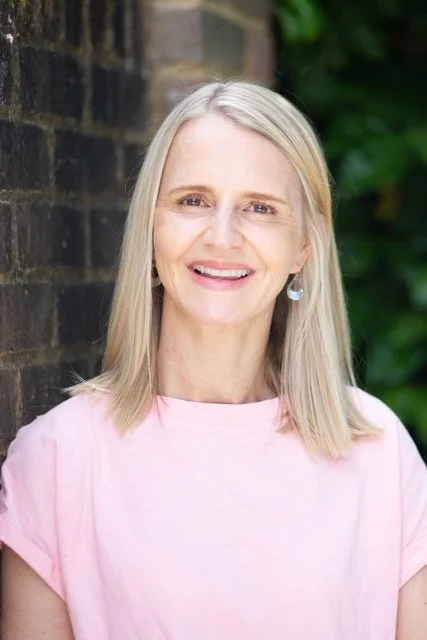 Claire Napier, smiling, wearing a light pink top and earrings, standing outdoors with greenery and a brick wall in the background.
