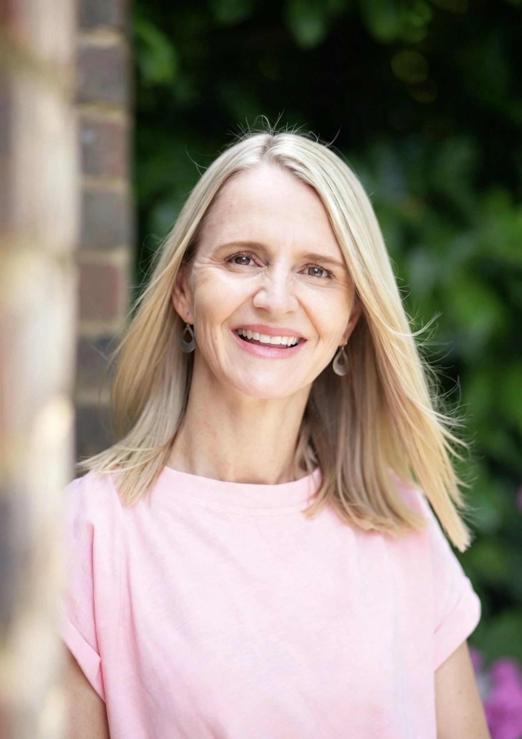Claire Napier standing outdoors against a background of green foliage and a brick wall.