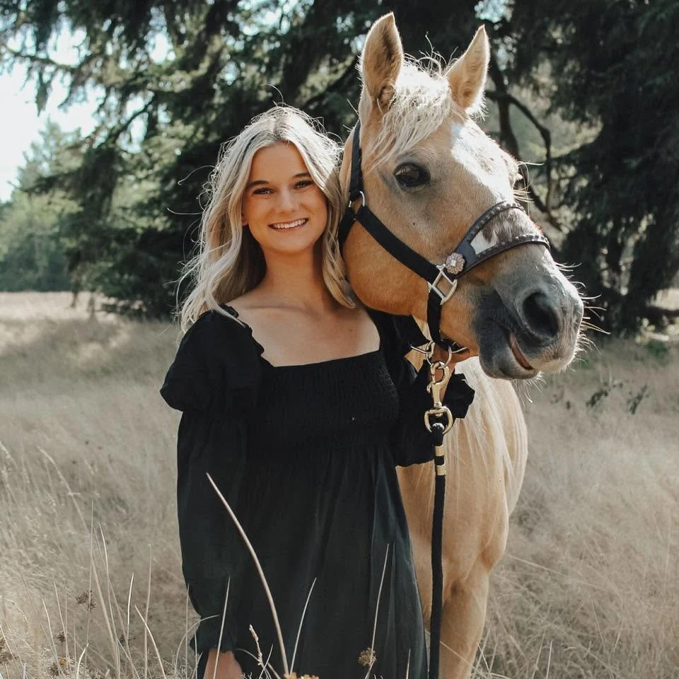 A young woman with blonde hair smiling and wearing a black dress standing next to a tan horse in an outdoor field with trees in the background.