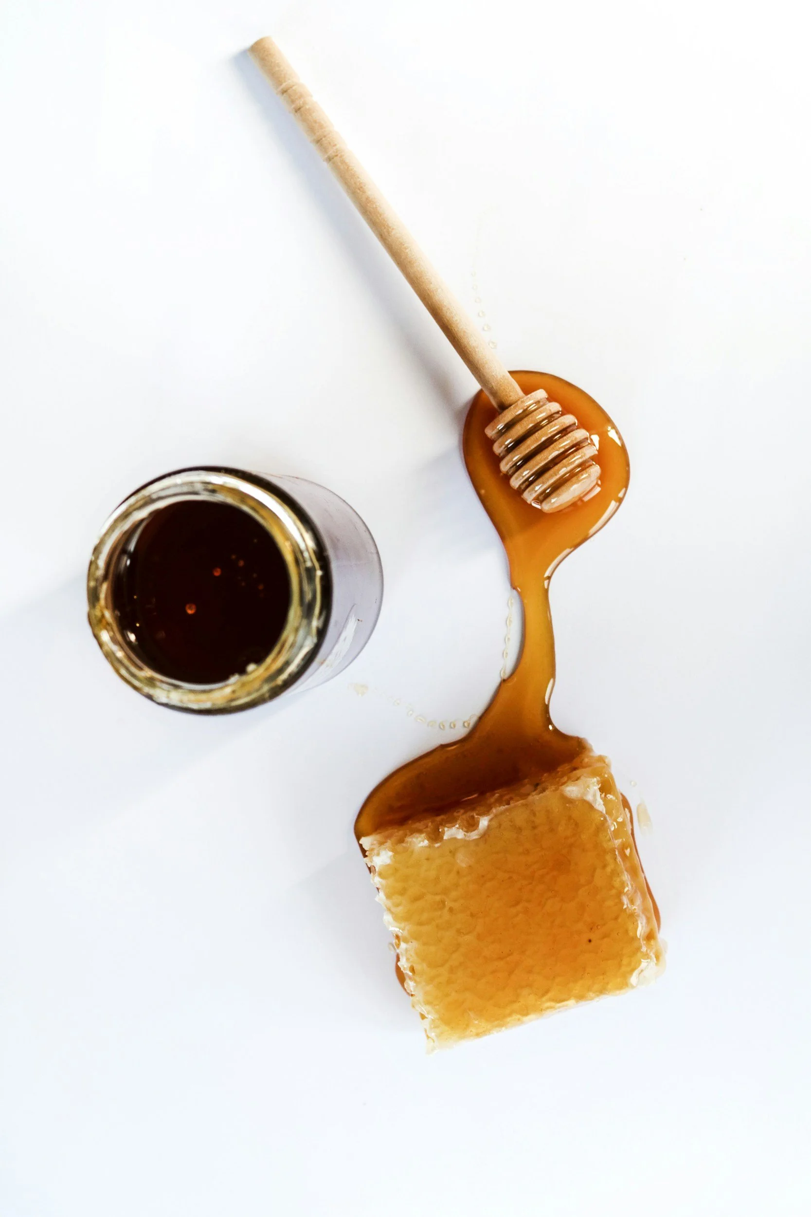 A jar of honey, a honey dipper with honey on it, honey dripping onto a honeycomb and honey spread on a white surface.