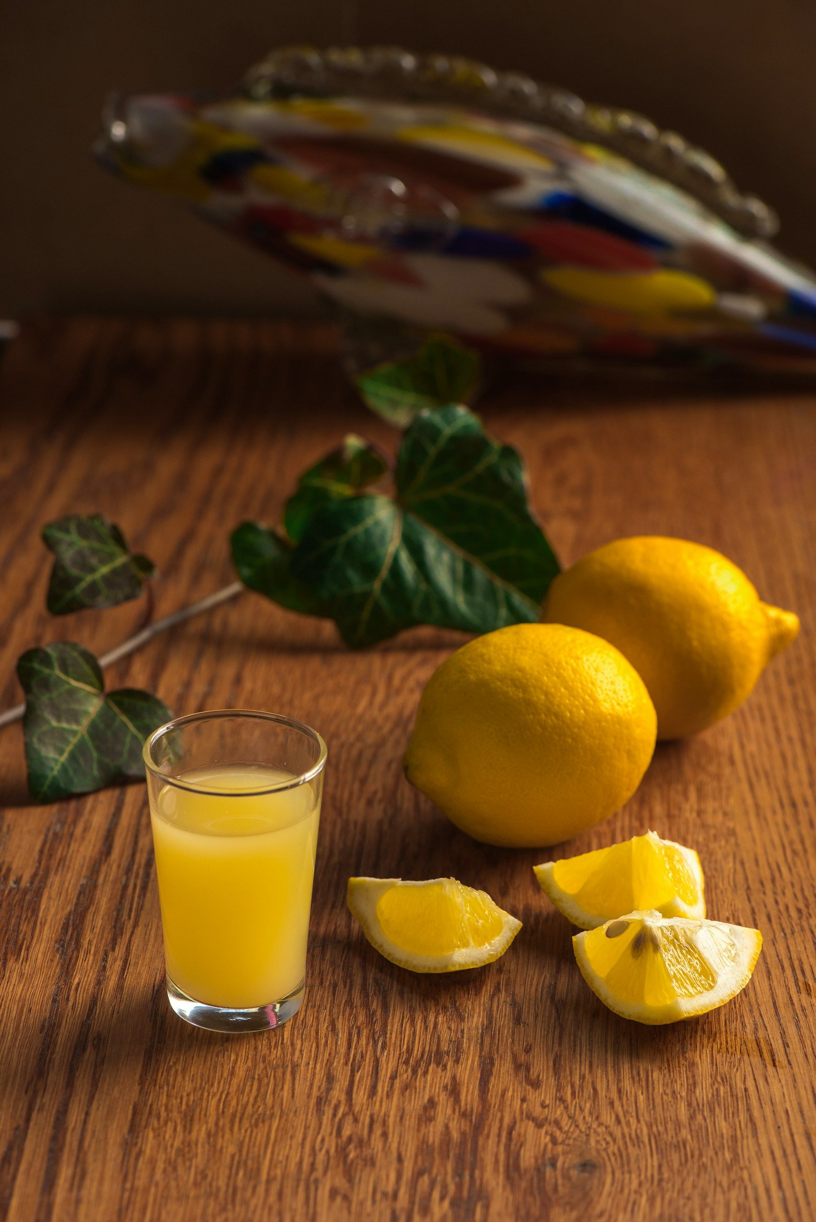 A glass of lemon juice, two whole lemons, lemon wedges, and lemon slices on a wooden surface with a blurred background of a decorative bowl and some green leaves.