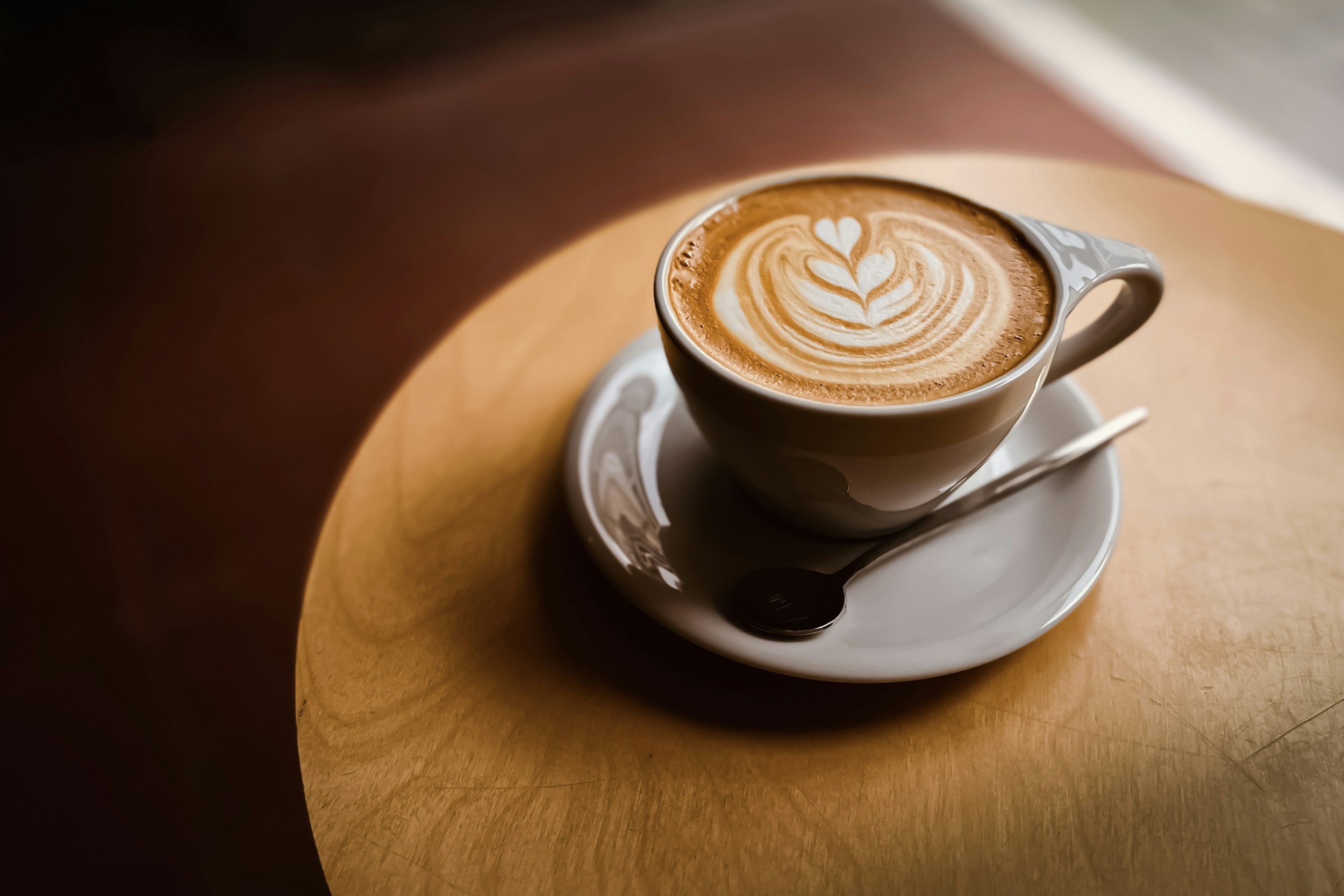 A ceramic cup of latte with latte art, placed on a saucer on a wooden table, with sunlight creating shadows.