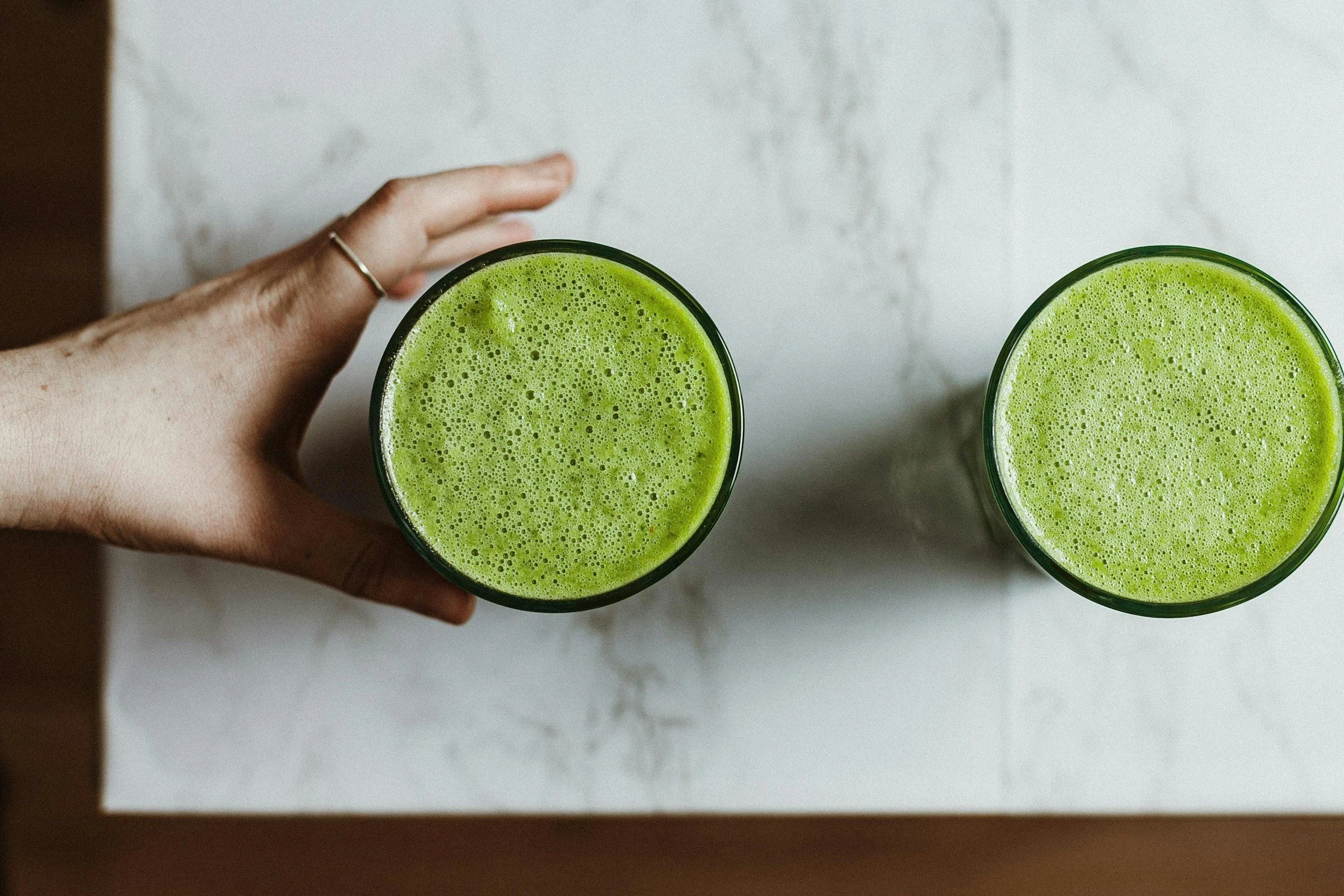 A top view of two glasses filled with green juice on a white marble surface, with a hand holding one glass on the left side of the image.