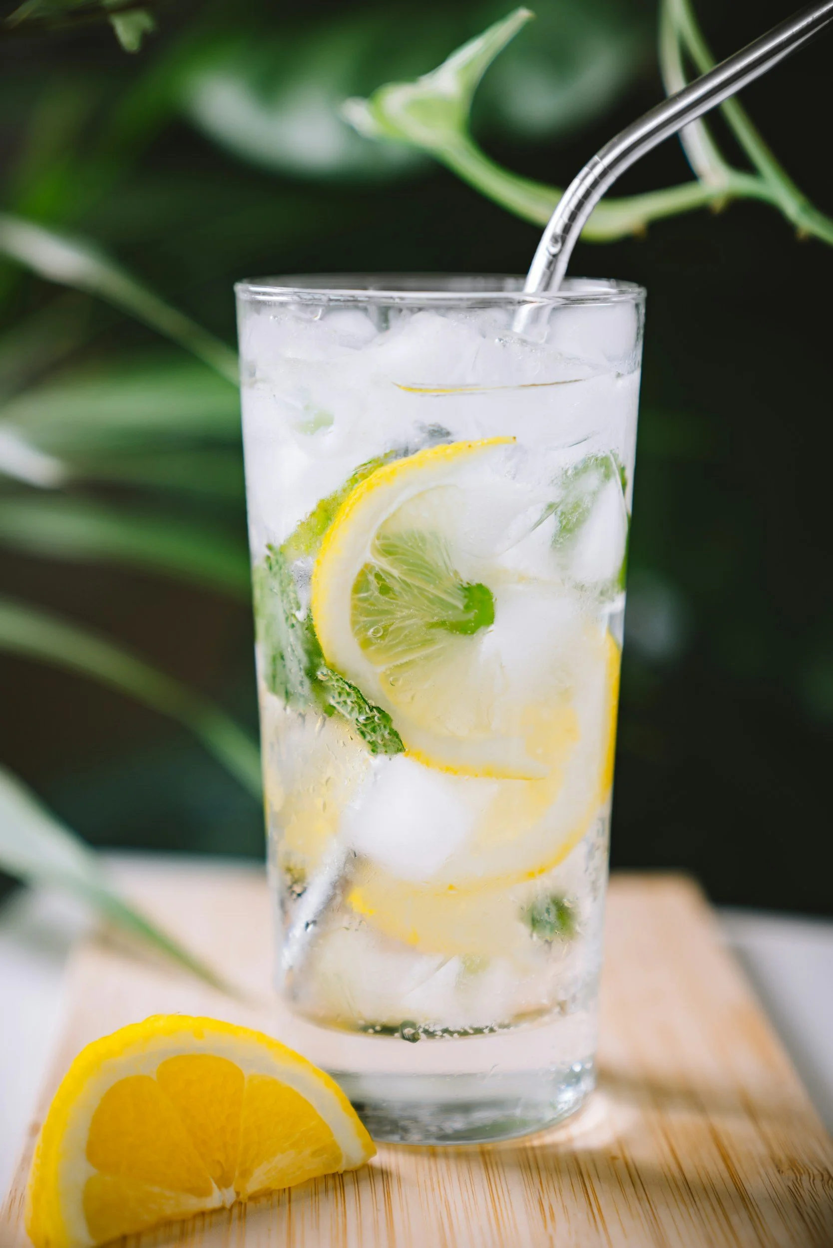 A tall glass of lemon and cucumber infused water with ice, with lemon slices and cucumber slices inside, placed on a wooden surface with a backdrop of green leaves.