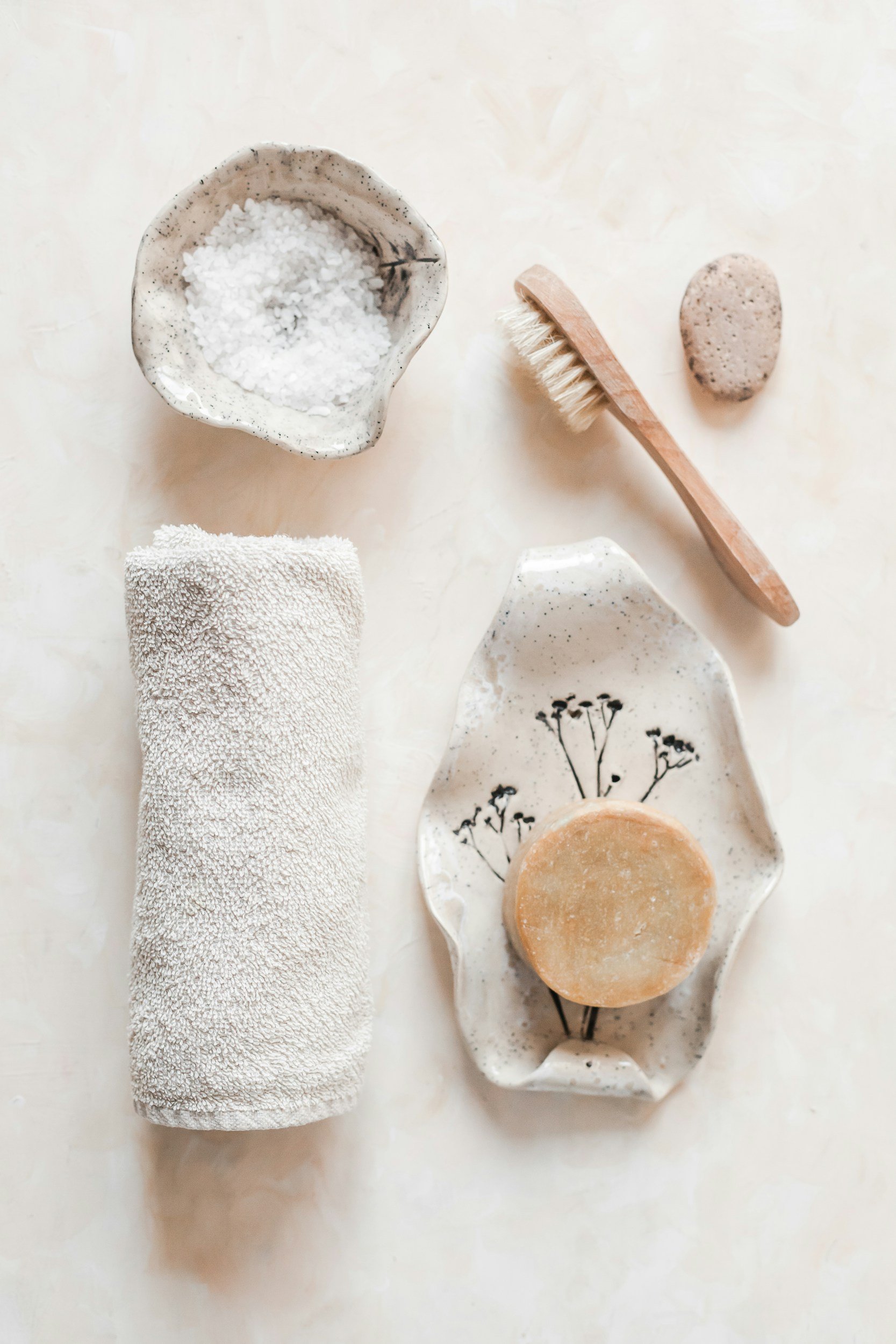 A spa setup with a rolled white towel, a white dish with black speckles containing coarse salt, a wooden body brush, a stone, a dish with black botanical sketches, and a round bar of soap on a light-colored surface.