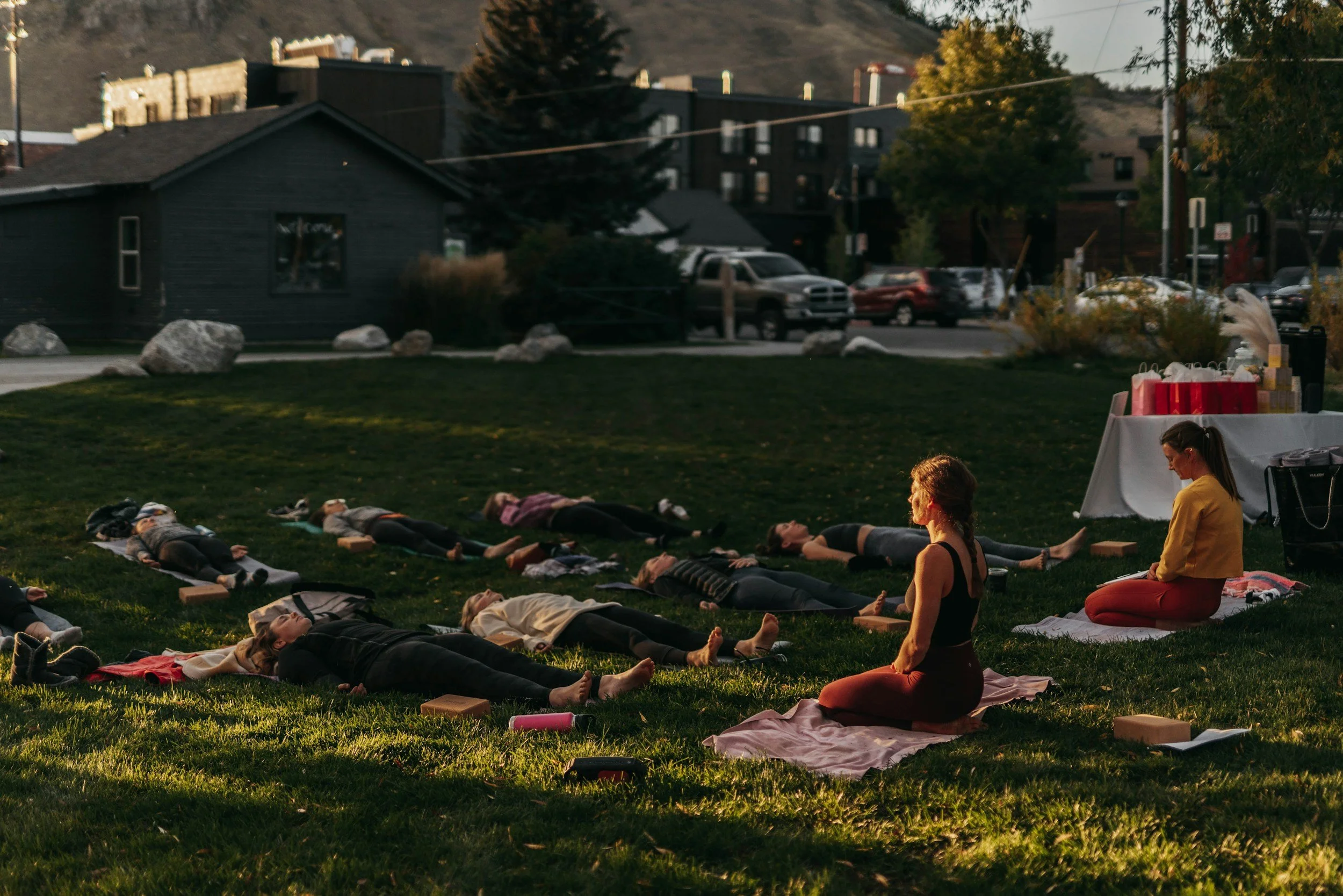 People participating in a yoga or meditation session outdoors on a grassy area in the evening, lying on mats with some sitting up nearby. There are rocks on the grass and a table with gift bags or decorations, with parked cars and buildings in the background.