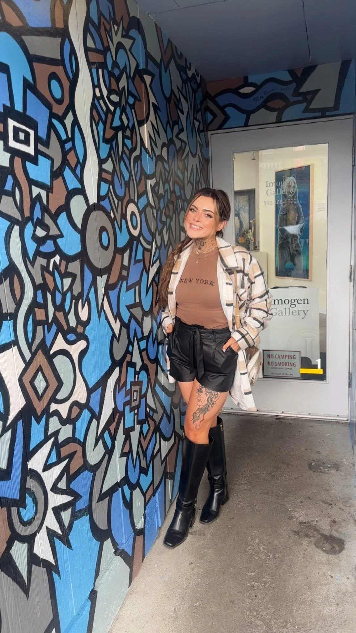 A young woman with long hair, tattoos, and a tan shirt that reads 'New York' stands smiling beside a large colorful mural on a wall. She is wearing a plaid coat, black shorts with a waist tie, and tall black boots. Behind her, a glass door has the sign 'Imogen Gallery' and contact information.