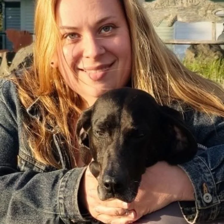 A woman with long red hair making a playful face and holding a black dog with dark eyes. The woman is wearing a denim jacket, and they are outdoors with a building and greenery in the background.