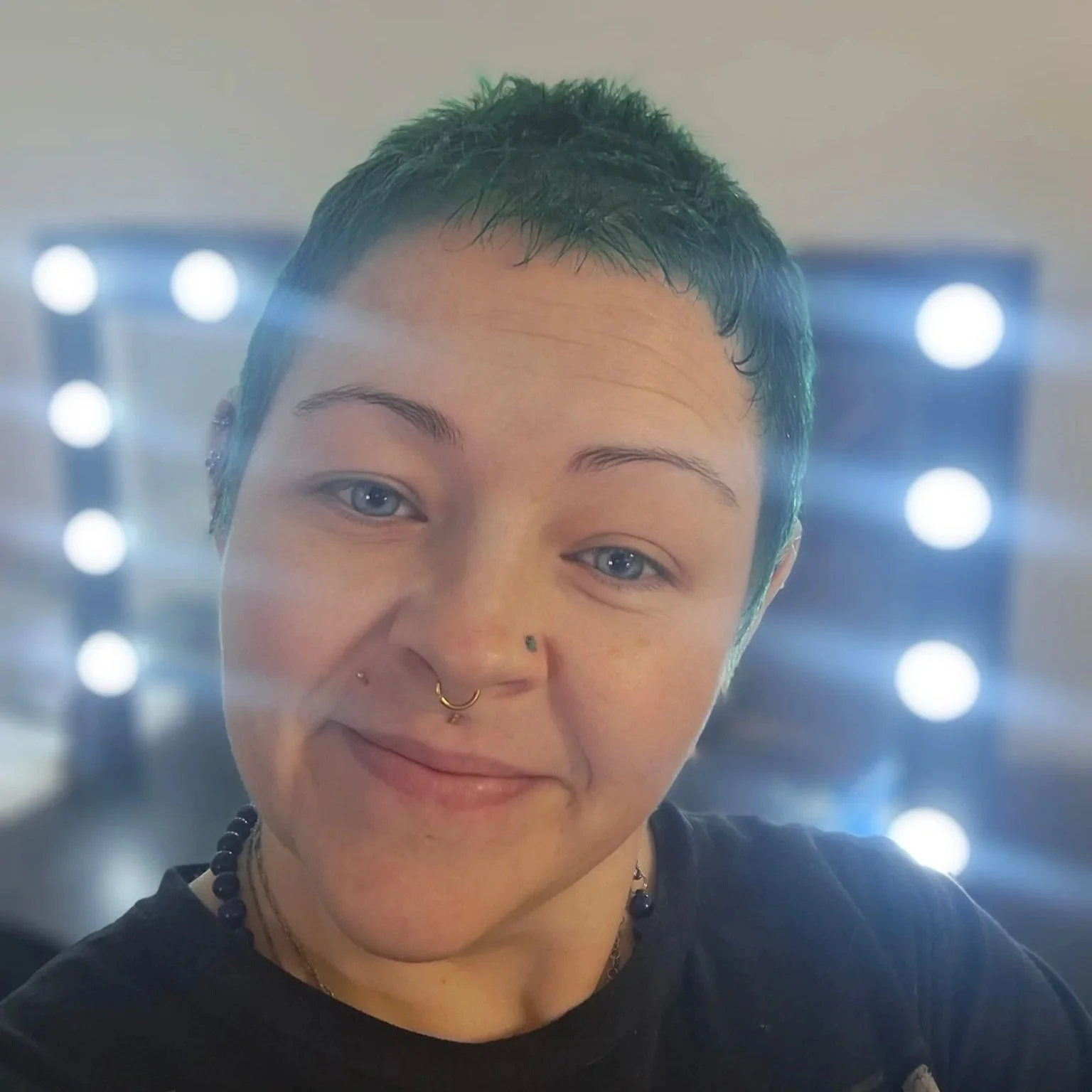 A person with short dark hair, blue eyes, and multiple facial piercings, smiling with a background of bright, circular lights.