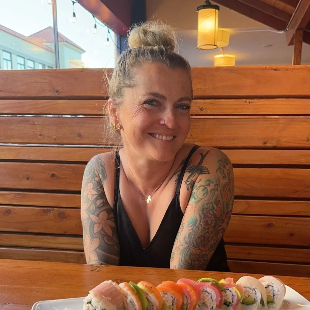 A smiling woman with tattoos on her arms, wearing a black tank top, sitting at a wooden table with a platter of assorted sushi in front of her in a restaurant with wooden decor and warm lighting.