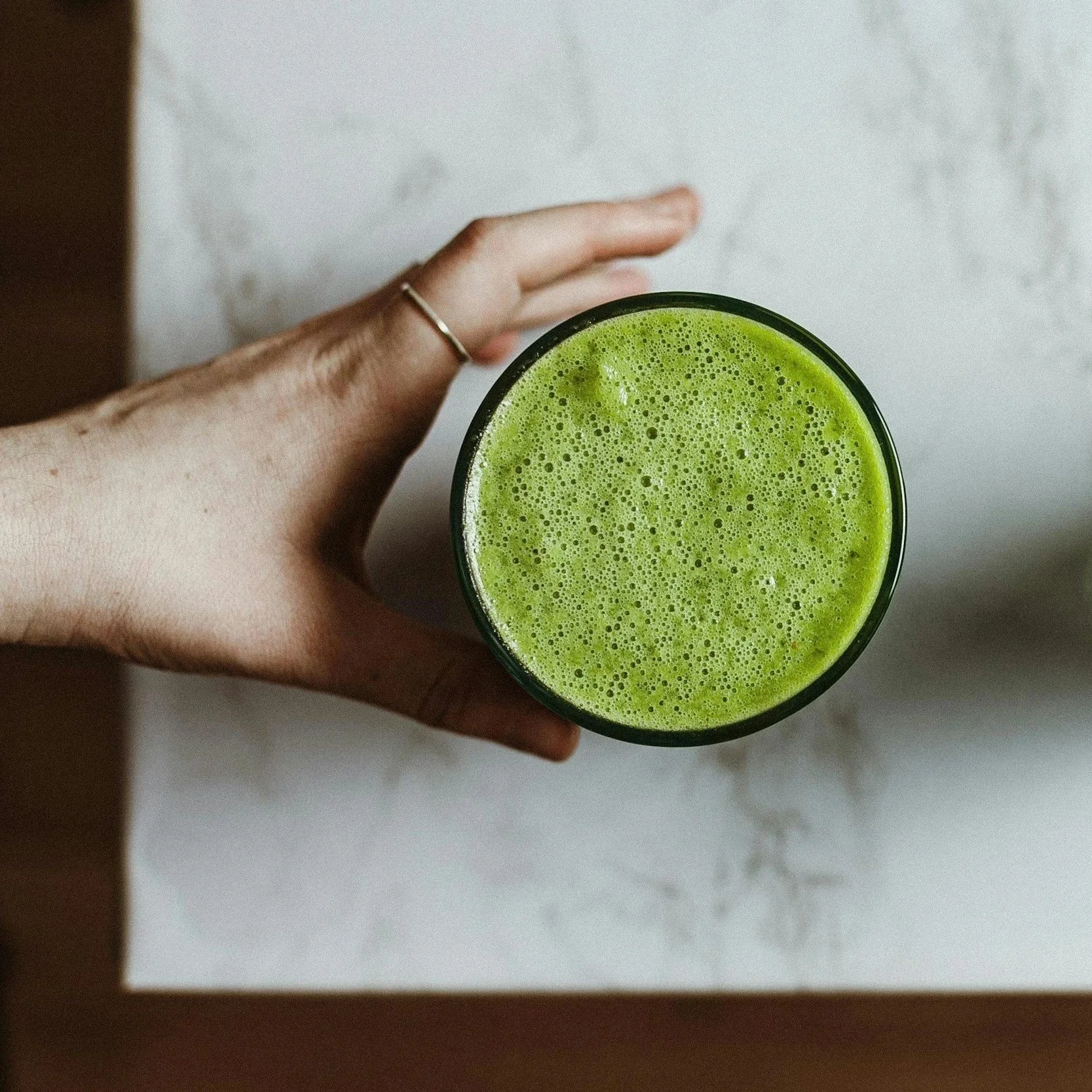 Top view of a glass filled with a bright green smoothie or juice, held by a person's hand with a small ring on the ring finger, against a textured white surface.