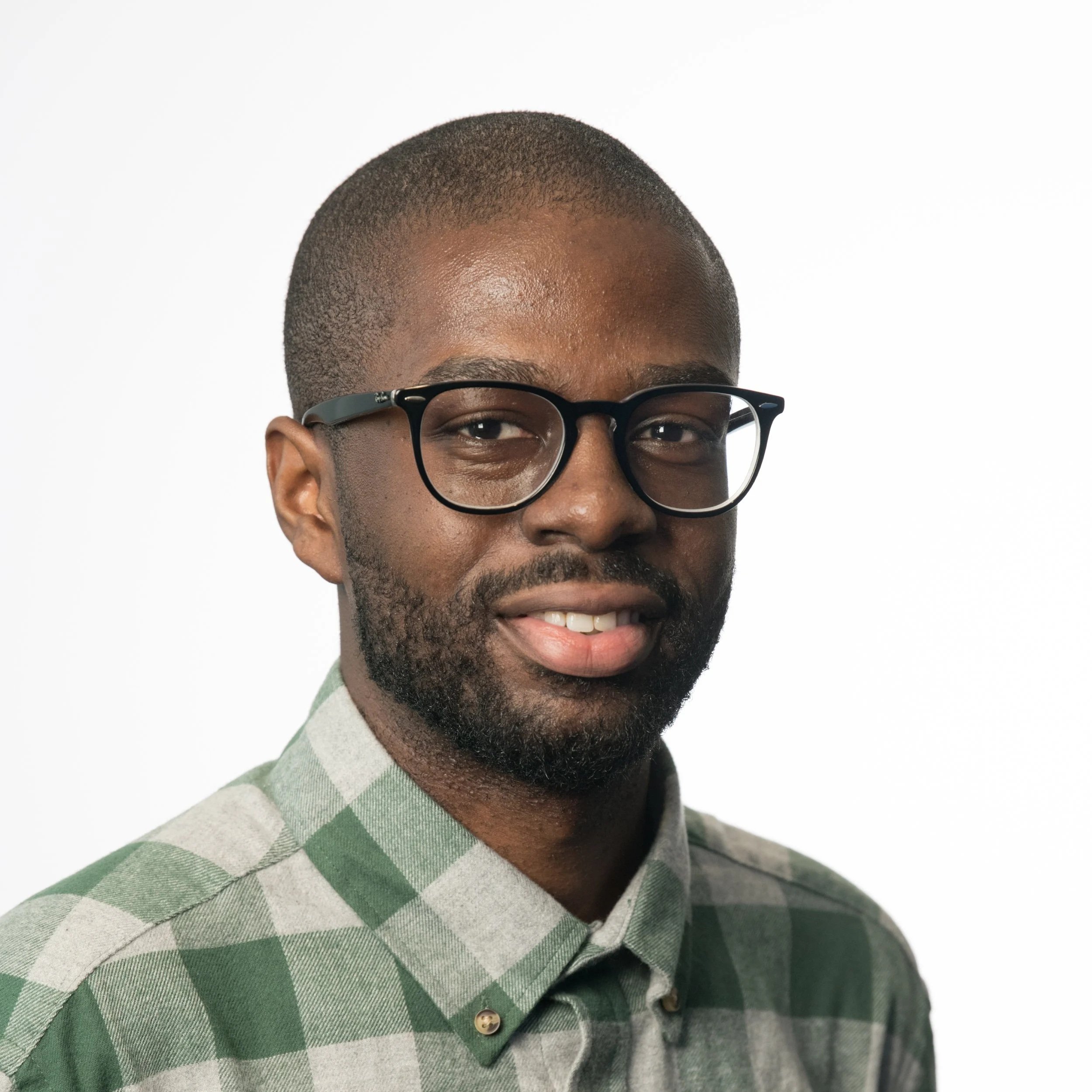 Close-up of a man's (Anthony K. McLean) face, wearing black glasses, with short hair and a beard, dressed in a checkered green and gray shirt, against a plain white background.