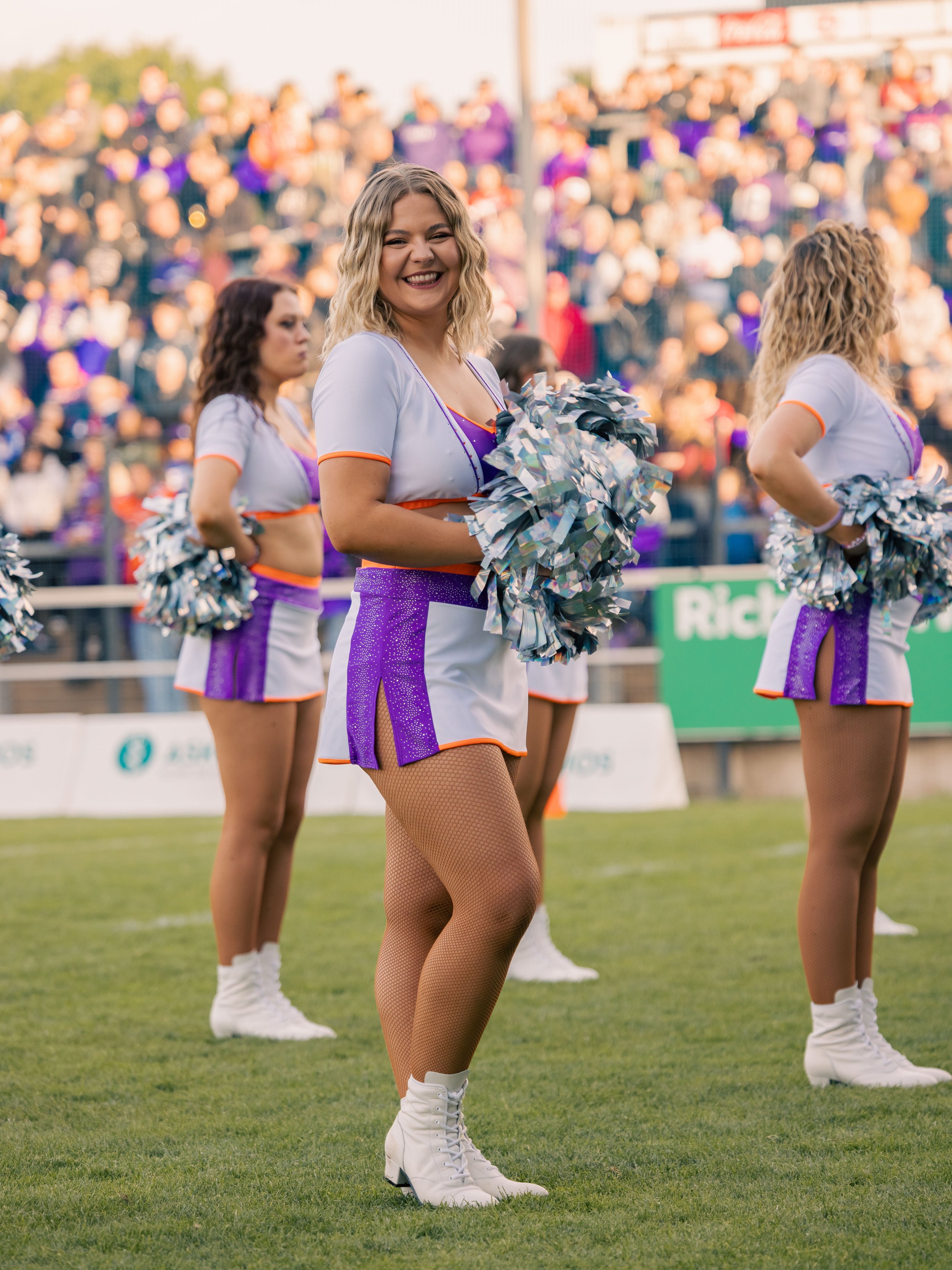 Tanzgruppe im Cheerleader-Outfit bei einem Sportevent, posieren auf dem Spielfeld, mit Zuschauern im Hintergrund.