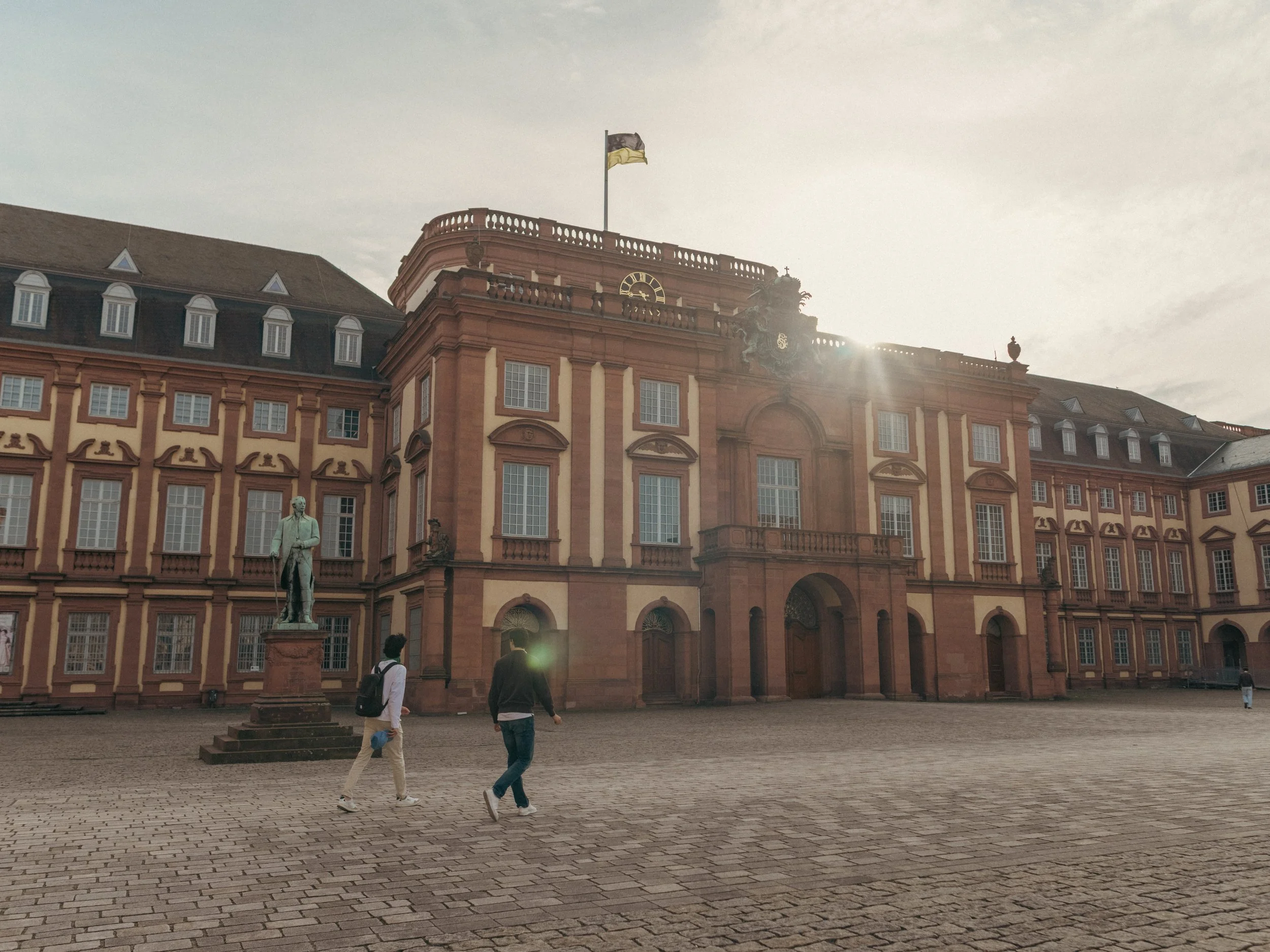 Das Mannheimer Schloss mit roten und weißen Fassaden, mehreren Fenstern und einem Turm. Zwei Menschen sitzen im Vordergrund, im Himmel weht eine Flagge. Bild für das Stadtmarketing Mannheim.