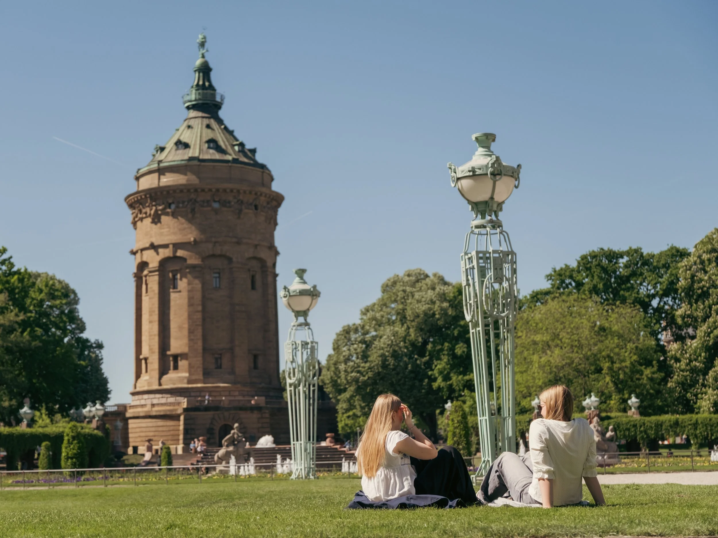 Zwei junge Frauen sitzen auf dem Rasen in einem Park, im Hintergrund der Mannheimer Wasserturm und schöne Bäume, blauer Himmel. Bild für das Stadtmarketing Mannheim.
