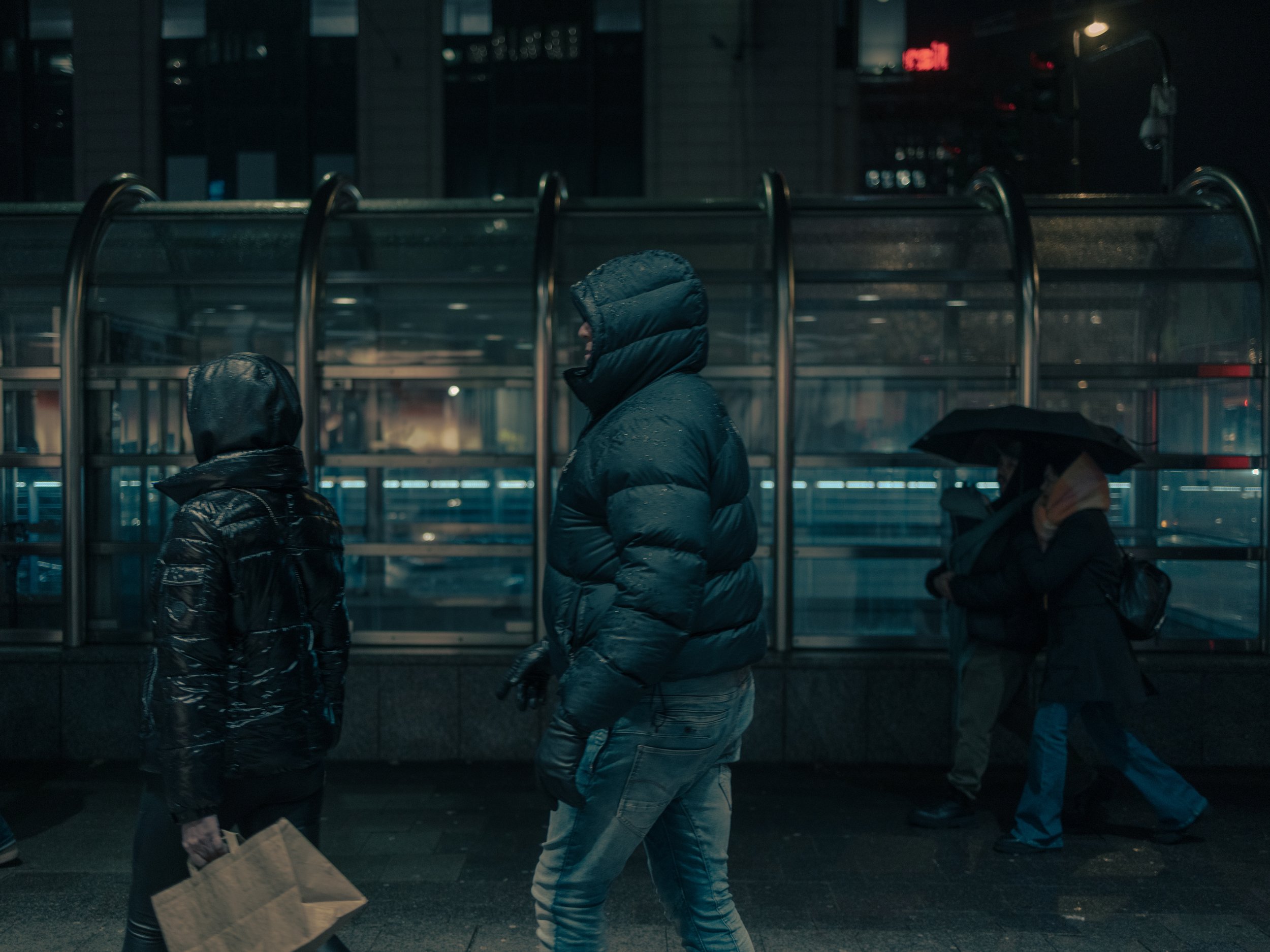 Menschen in dunkler Oberbekleidung stehen bei Nacht an einer Bushaltestelle in einer Stadt, im Hintergrund leuchtende Lichter und ein Gebäude. Streetphotography in Düsseldorf.