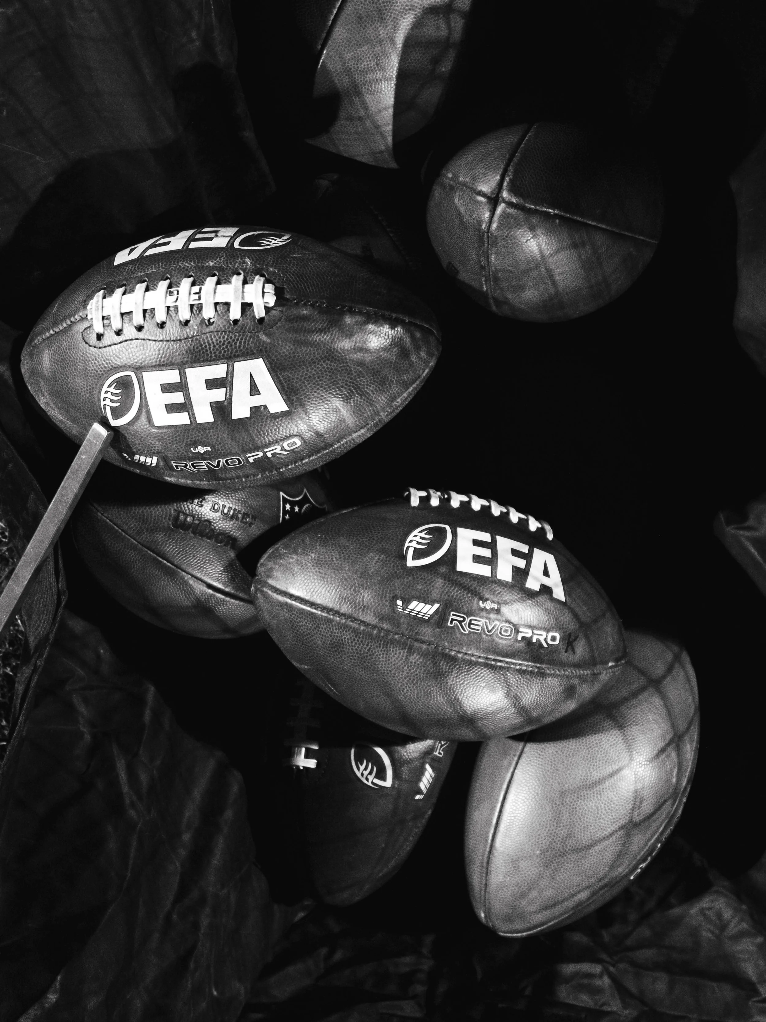 Mehrere schwarze American Footballs mit weißen Aufschriften und Logos, einige mit weißen Schnürsenkeln, liegen auf schwarzem Kunststoff. Teil der European Football Alliance.
