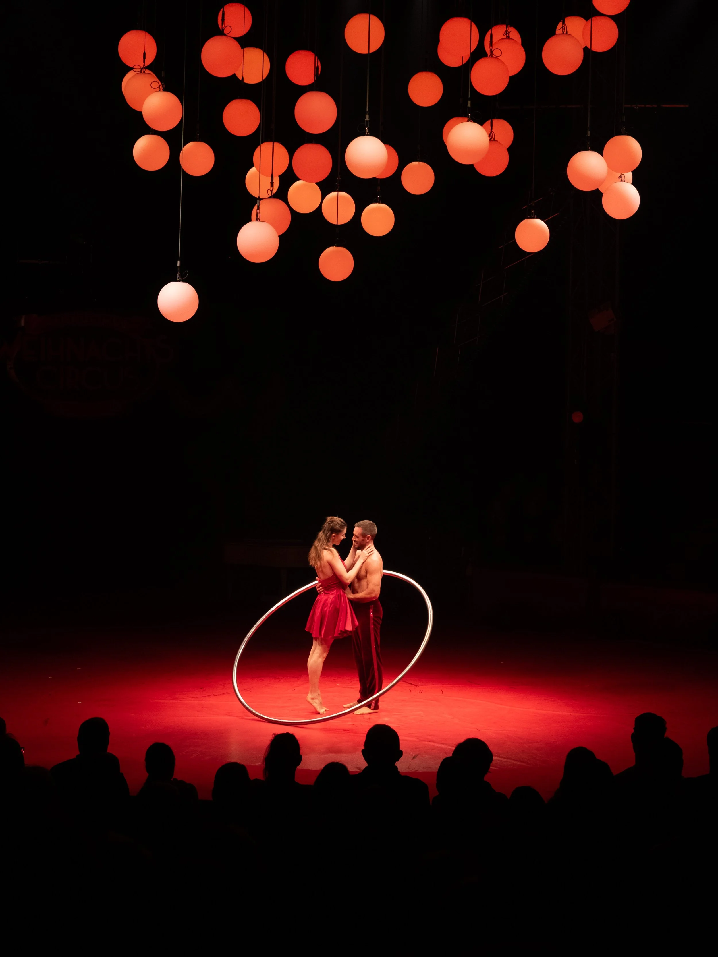 Zwei Akrobaten tanzen in der Mitte einer Bühne des Mannheimer Weihnachtszirkus, umgeben von roten Lichtern und dunklem Hintergrund, Zuschauer im Vordergrund. Event-Fotografie in Mannheim.
