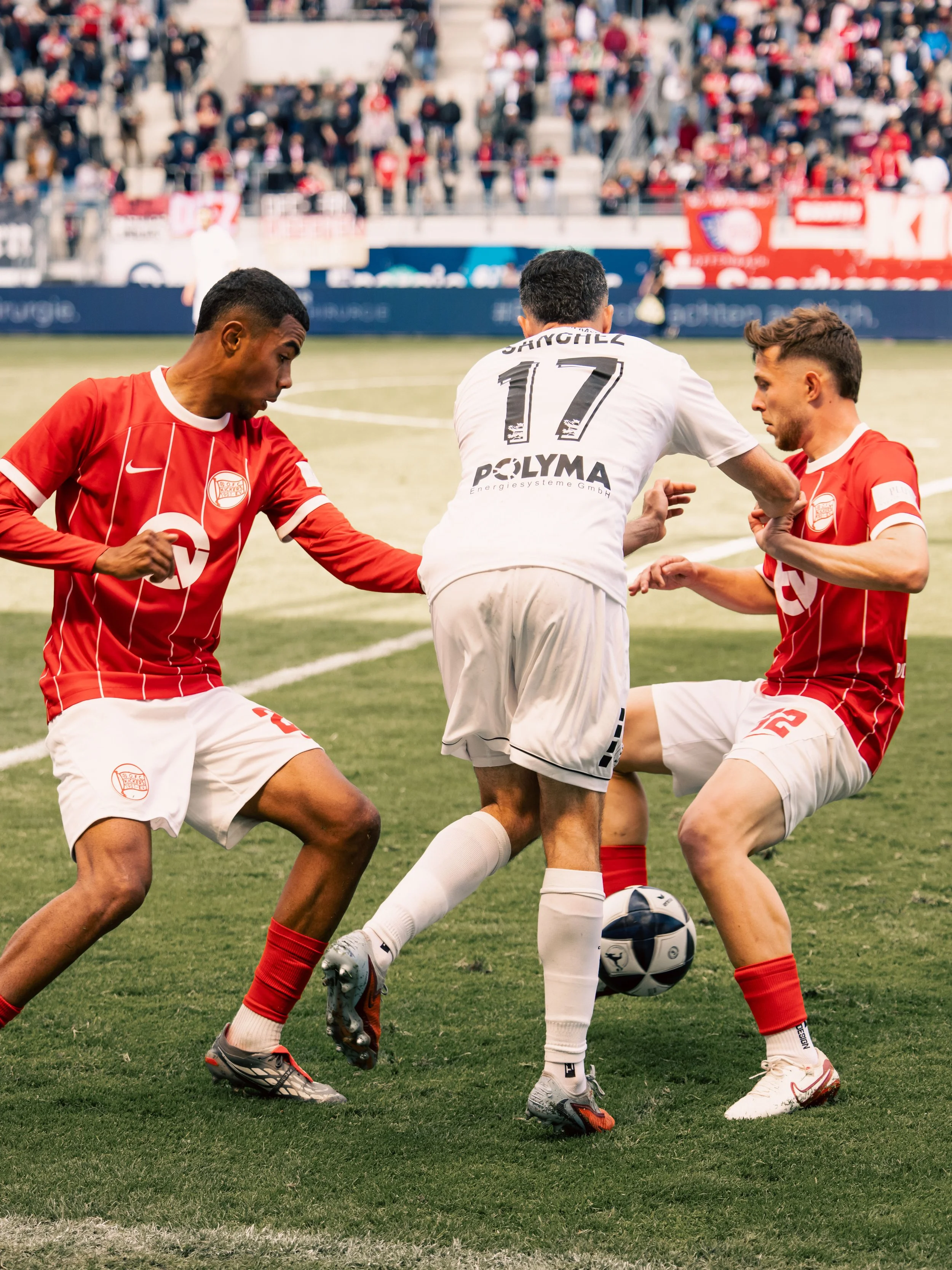 Fußballspieler kämpfen um den Ball auf dem Spielfeld während eines Spiels, im Hintergrund stehende Zuschauermenge im Stadion.