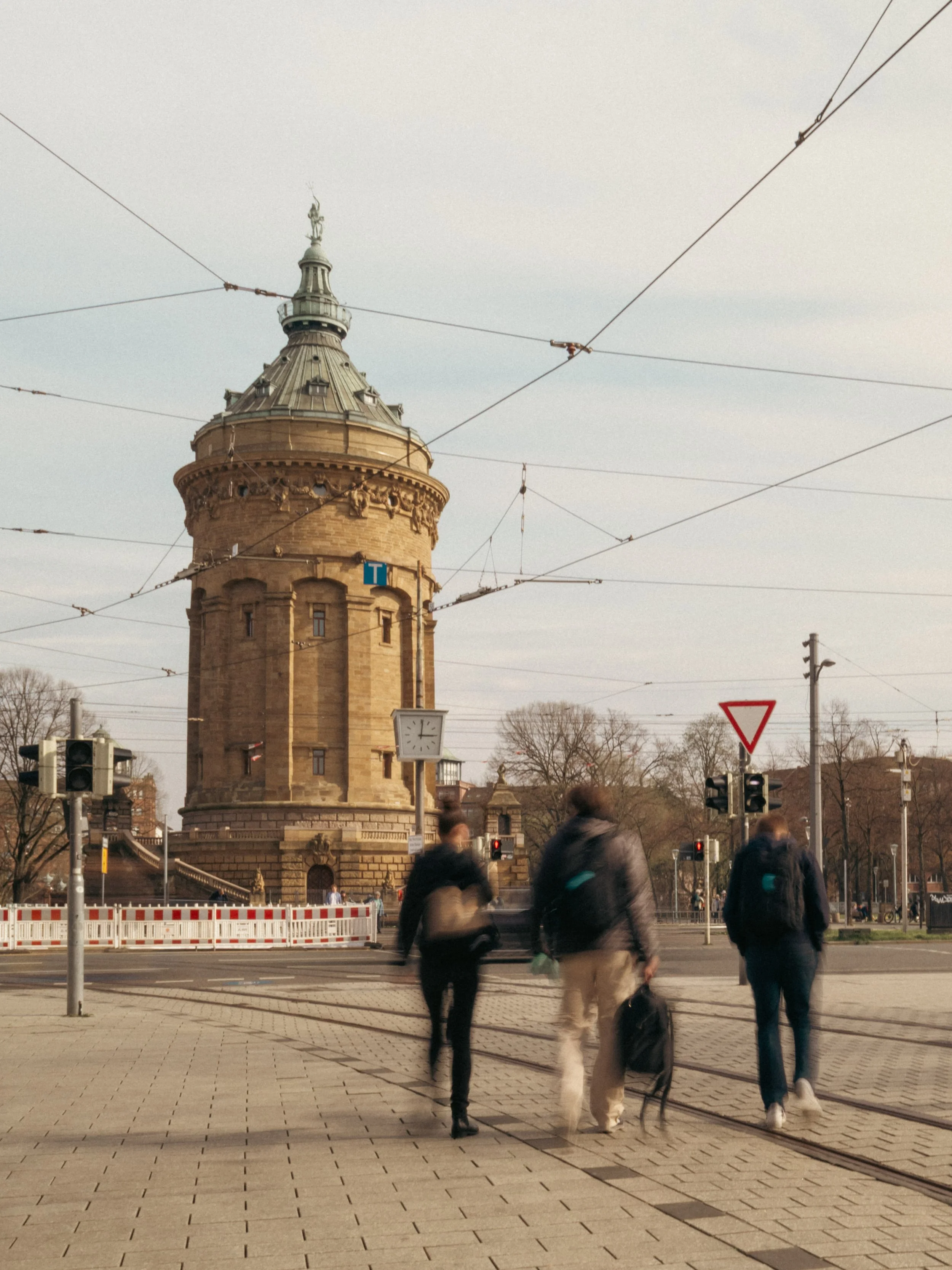Blick auf den Mannheim Wasserturm, darüber hängen Kabel der Straßenbahnen, im Vordergrund Menschen, die die Straße überqueren. Bild für das Stadtmarketing Mannheim.