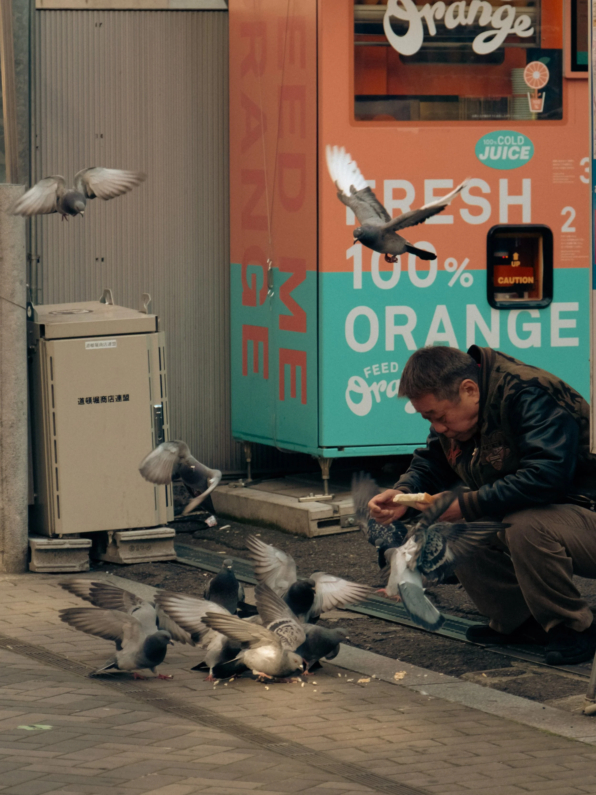Ein Mann füttert Tauben auf einem Bürgersteig an einer Ecke. Im Hintergrund sind ein orangefarbener Getränkeautomat und eine Wand. Tauben fliegen um den Mann herum, während er sie füttert. Street-Photography in Japan.
