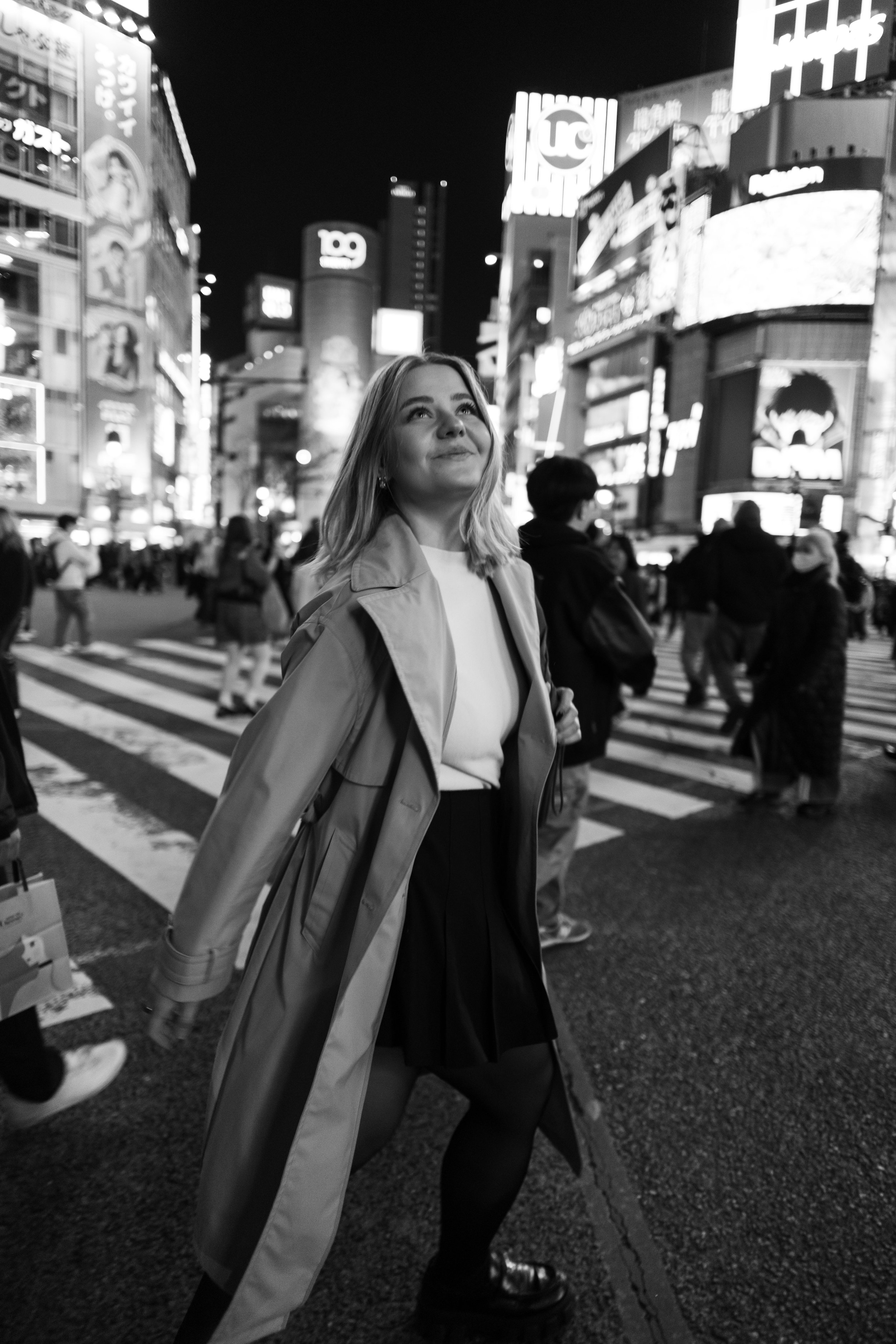 Eine lachende junge Frau steht auf einer belebten Straße auf der Shibuya-Kreuzung in einer Stadt bei Nacht, umgeben von hell beleuchteten Werbetafeln und Menschen.