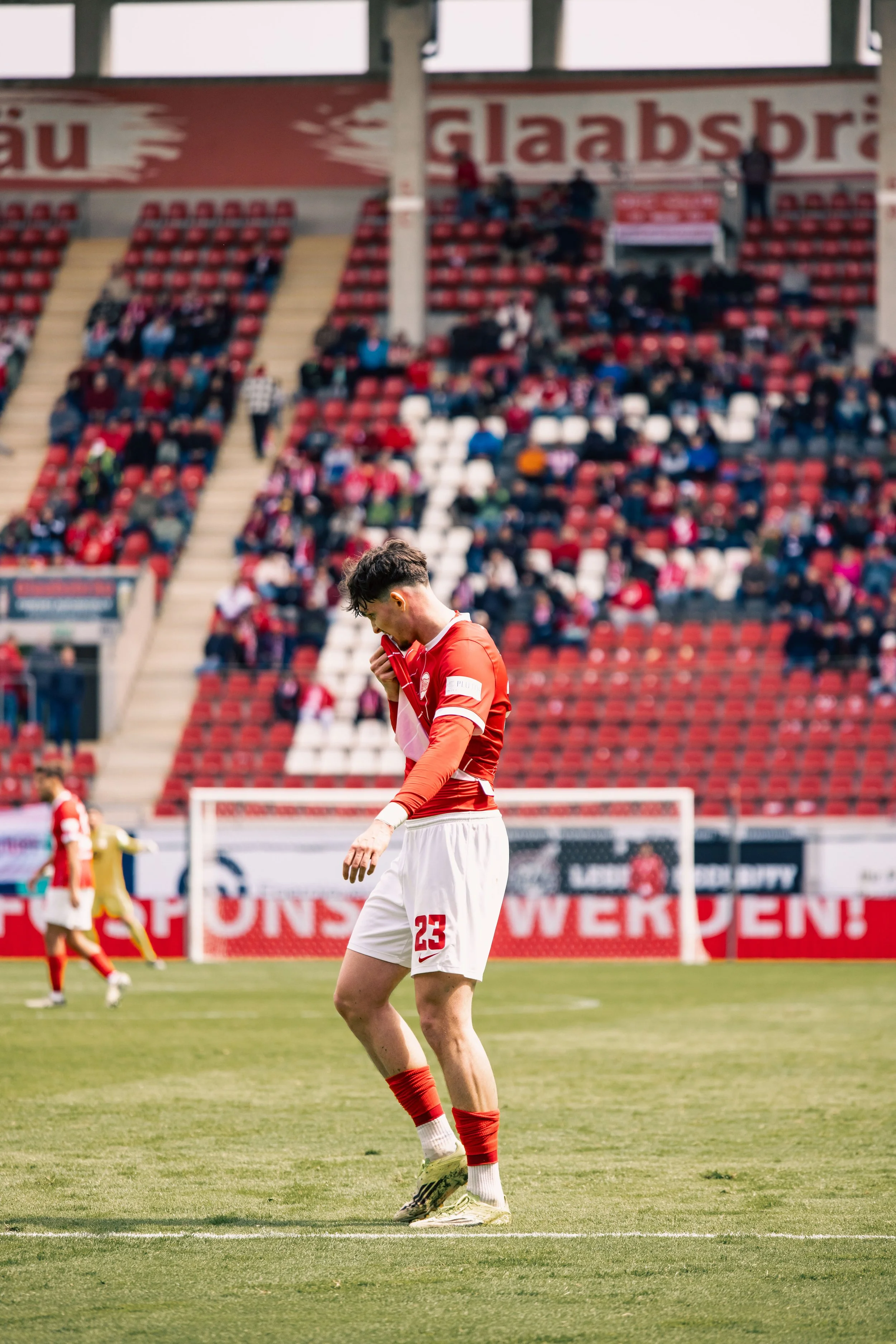 Fußballspieler in rotem und weißem Trikot auf dem Spielfeld, Blick nach unten, inmitten eines Spiels mit Zuschauern im Hintergrund.