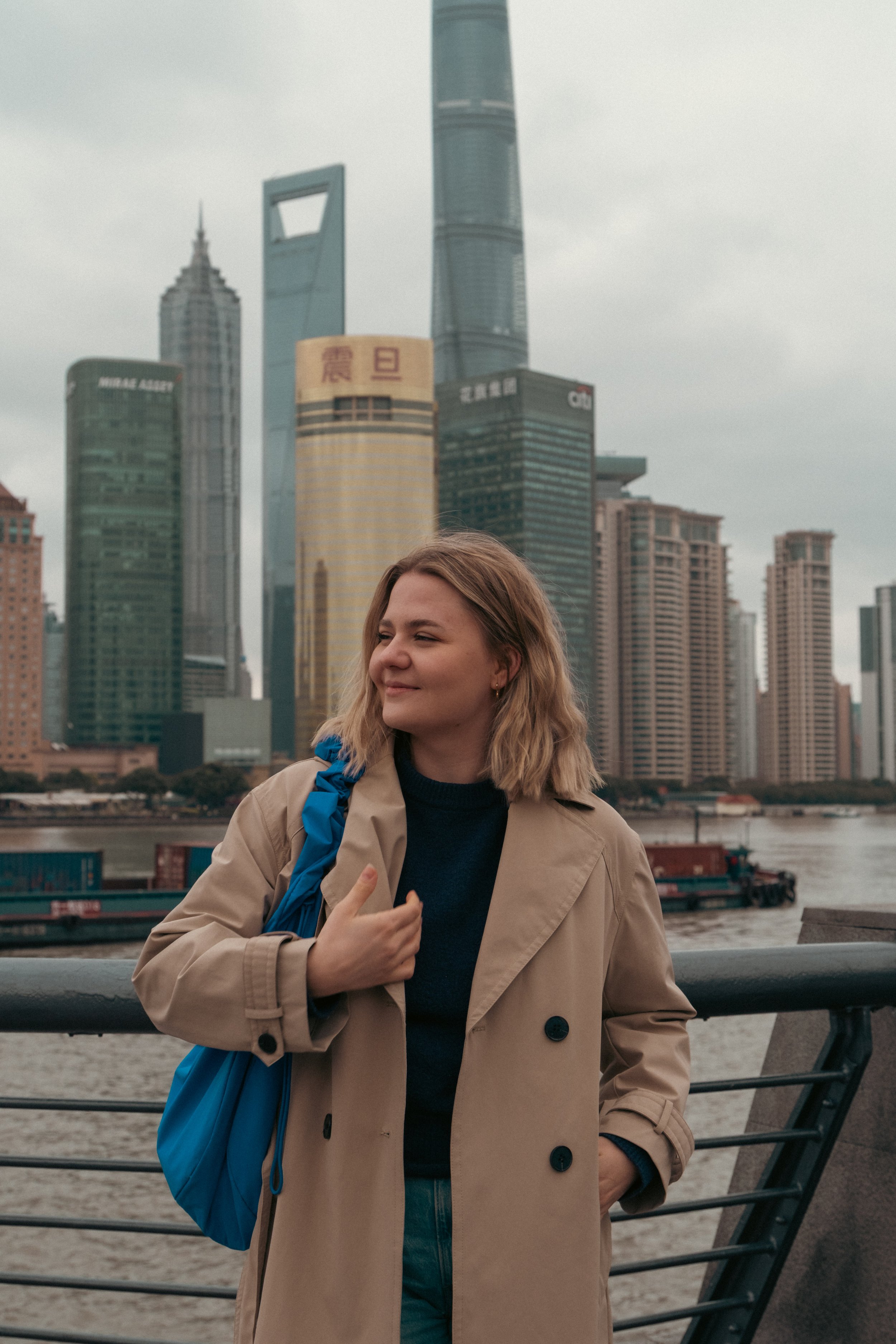 Junge Frau mit blondem Haar, Hellbraune Trenchcoat und blauer Tasche steht am Fluss in Shanghai, im Hintergrund die Skyline mit den Wolkenkratzern Jin Mao Tower, Shanghai World Financial Center und Shanghai Tower bei bewölktem Himmel.