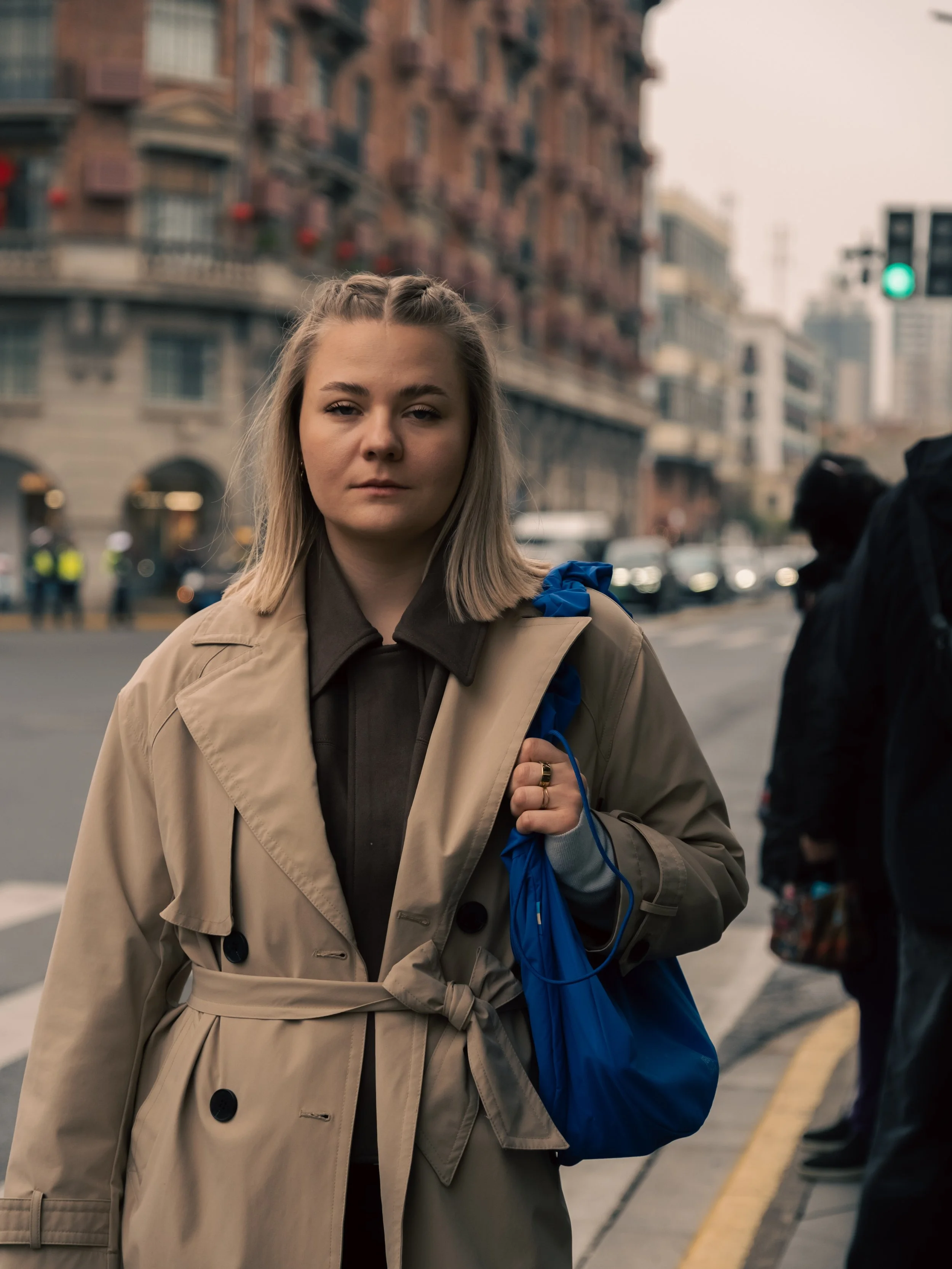Eine junge Frau mit blonden Haaren trägt einen beige Trenchcoat und hält eine blaue Tasche, während sie auf einer städtischen Straße in Shanghai, China, steht. Im Hintergrund sind Gebäude und Fußgänger zu sehen.
