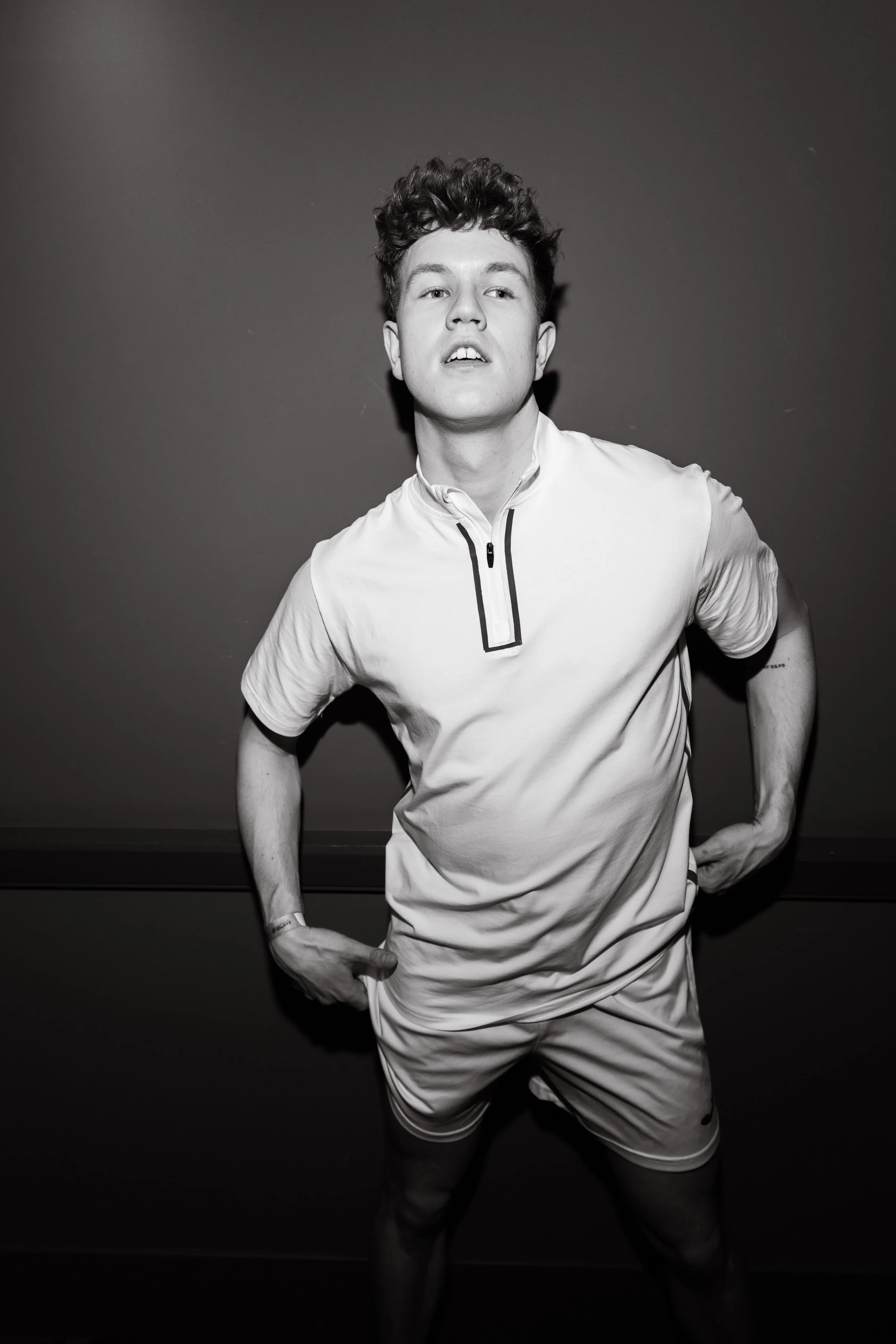 Schwarz-Weiß-Foto eines jungen Mannes, der ein sportliches Cheerleader-Outfit trägt, mit lockigem Haar, und in einer dynamischen Pose steht, die auf Energie und Selbstvertrauen hinweist.