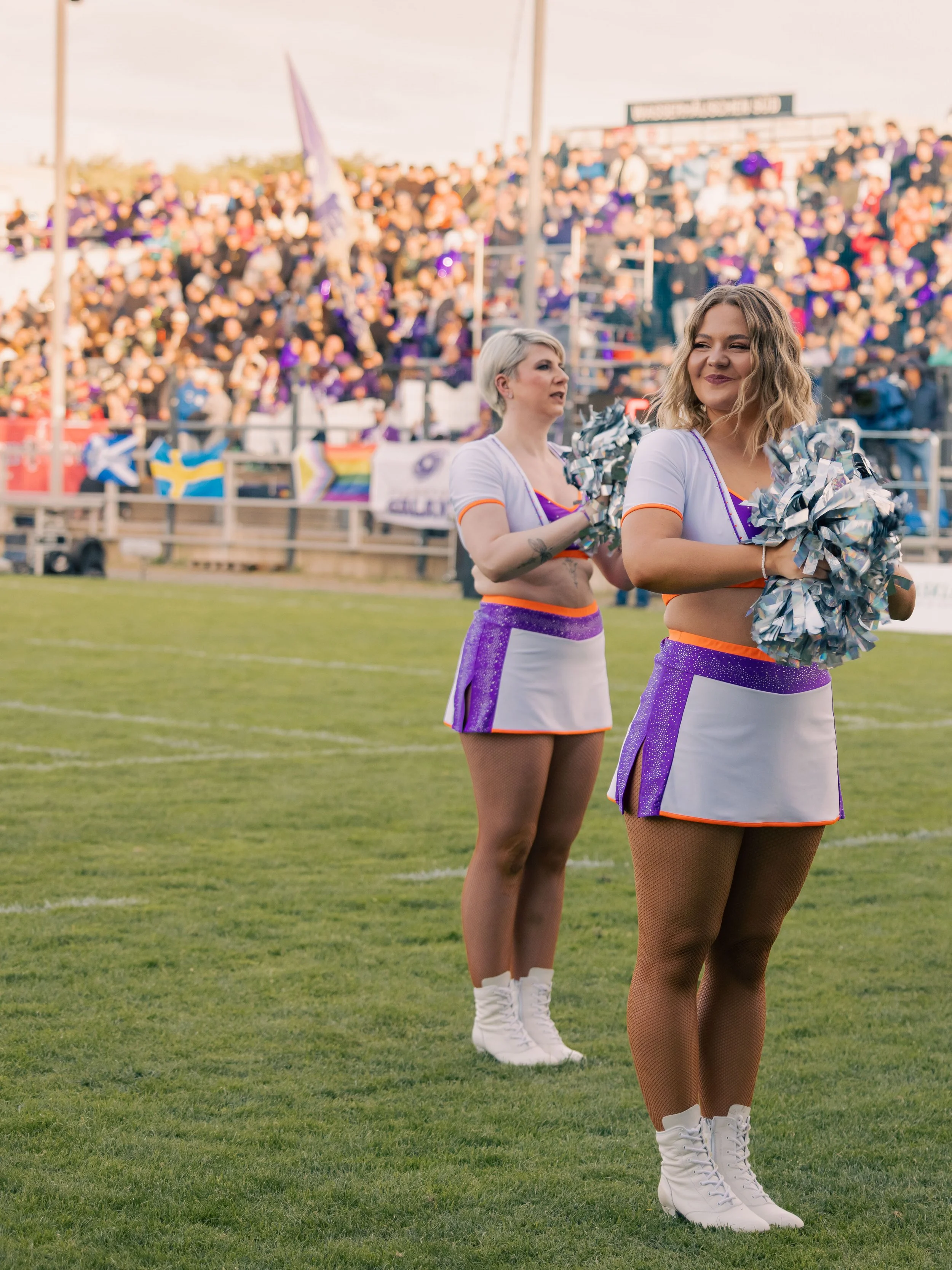 Zwei Cheerleaderinnen in weißen und lila-outfits mit Pompons auf einem Fußballfeld vor einer großen Menschenmenge im Stadion.