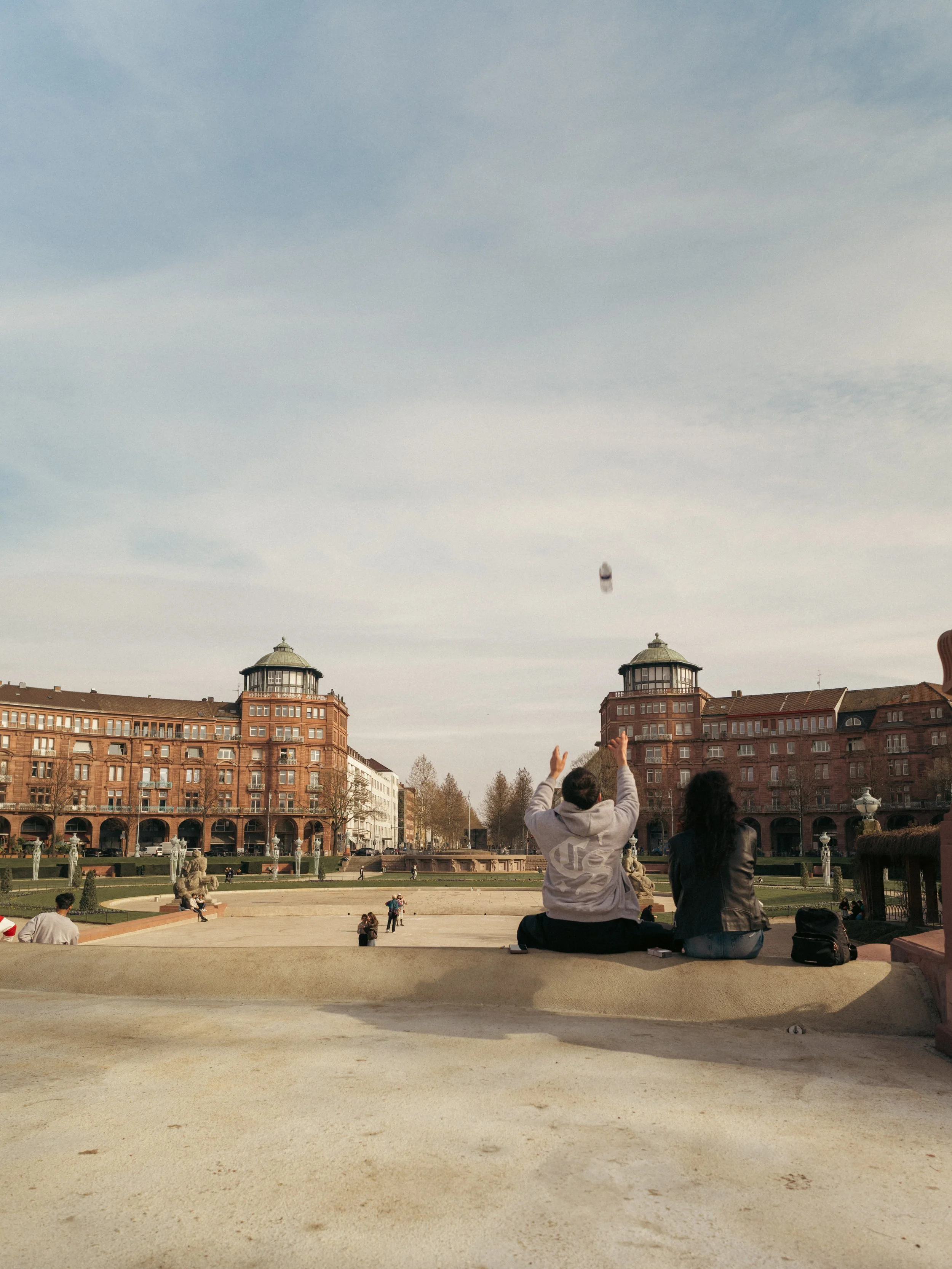 Menschen sitzen am Wasserturm in Mannheim in einem gepflegten Parkplatz mit Blick auf ein historisches Gebäude im Hintergrund, werfen eine Flasche nach oben. Bild für das Stadtmarketing Mannheim.